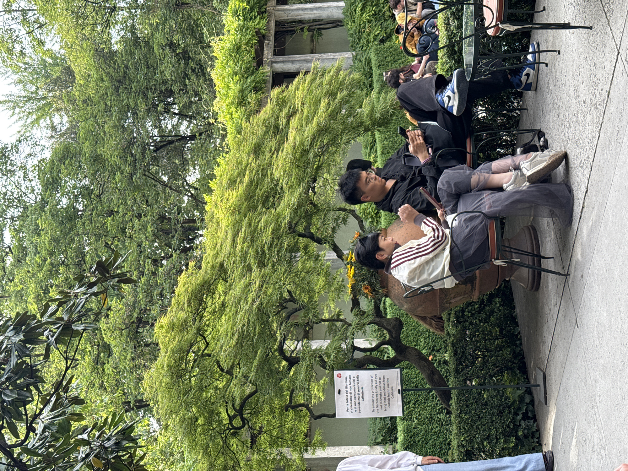 Here's a forensic description of the image:
**Setting:** An outdoor garden setting in Milan, Italy. The environment includes mature trees and hedges. There's a sign indicating rules regarding smoking and consumption of outside food and beverages. The sign is in both Italian and English, mentioning the "Cafetteria."
**Subjects:** Several individuals are present. Two people are seated at a table, seemingly engrossed in their phones. Other people are visible in the background, also seated at tables.
**Objects:** Tables and chairs are set up for patrons. A planter or vase is used as the base for a table. One of the chairs has a backpack hanging on it. There is a sign with instructions.
**Actions:** The people at the foreground table appear to be relaxing, using their phones. Other people are engaged in similar activities in the background.
**Interactions:** People are interacting with their phones. The main interaction is sitting down and relaxing in the garden setting.