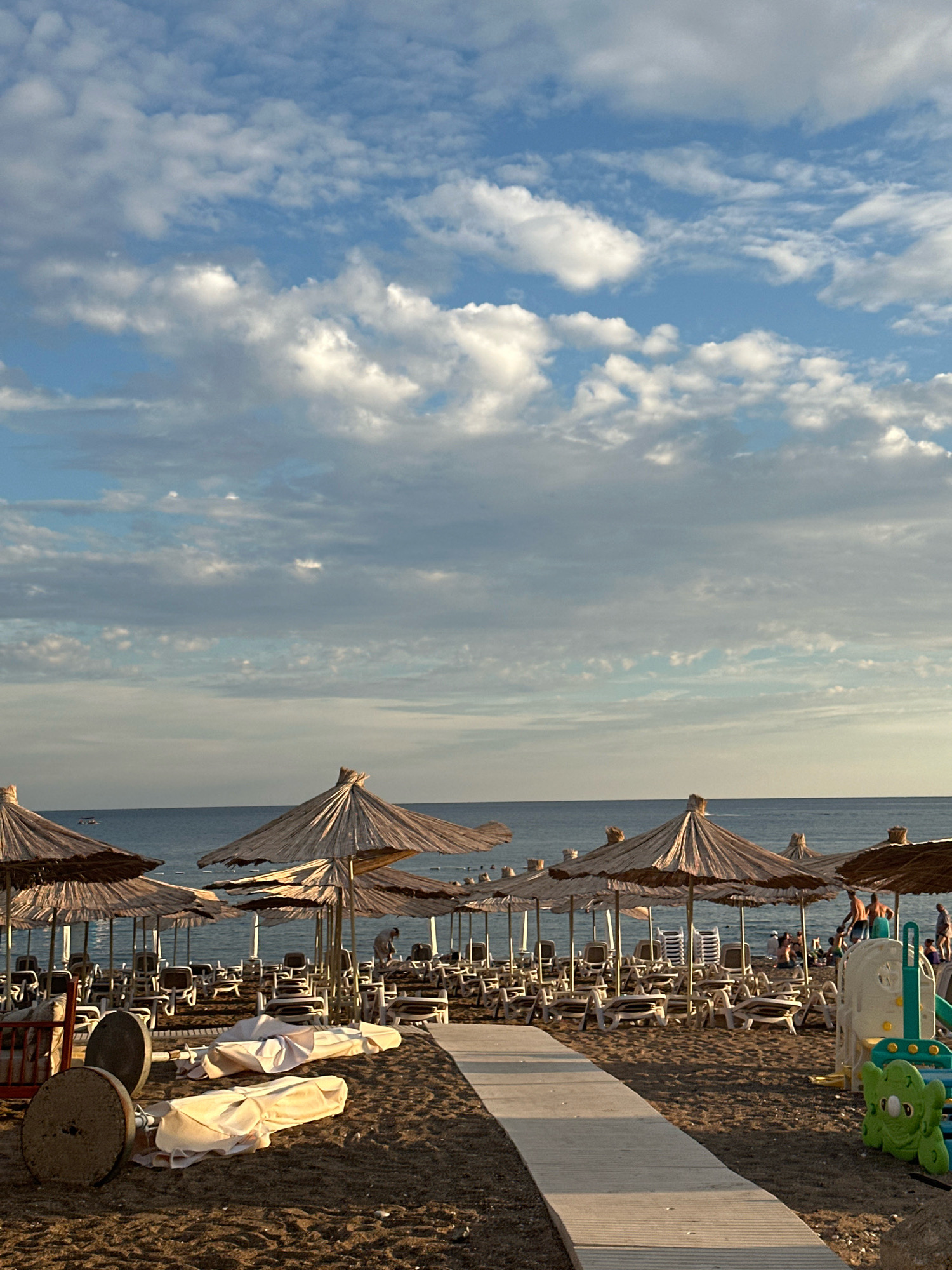 This is a medium-shot, eye-level, vertical photograph of a sandy beach in Boreti, Montenegro, on a cloudy day. The sky is filled with a mix of blue sky and white and gray clouds, with sunlight breaking through in some areas. The ocean is visible in the background, a calm, deep blue. In the foreground, a wooden walkway leads from the bottom of the frame towards the beach. To the left of the walkway, several beach umbrellas are folded and appear to be stored, with two large, round, metallic objects on stands nearby. Further back, rows of white lounge chairs are set up on the sand, mostly unoccupied. Several straw-covered beach umbrellas are open, providing shade over some of the chairs. A few people are visible in the distance, near the water's edge, some swimming or wading. To the right of the walkway, a small, colorful children's play structure, possibly a slide, is set up on the sand. The overall impression is a peaceful, end-of-day beach scene.