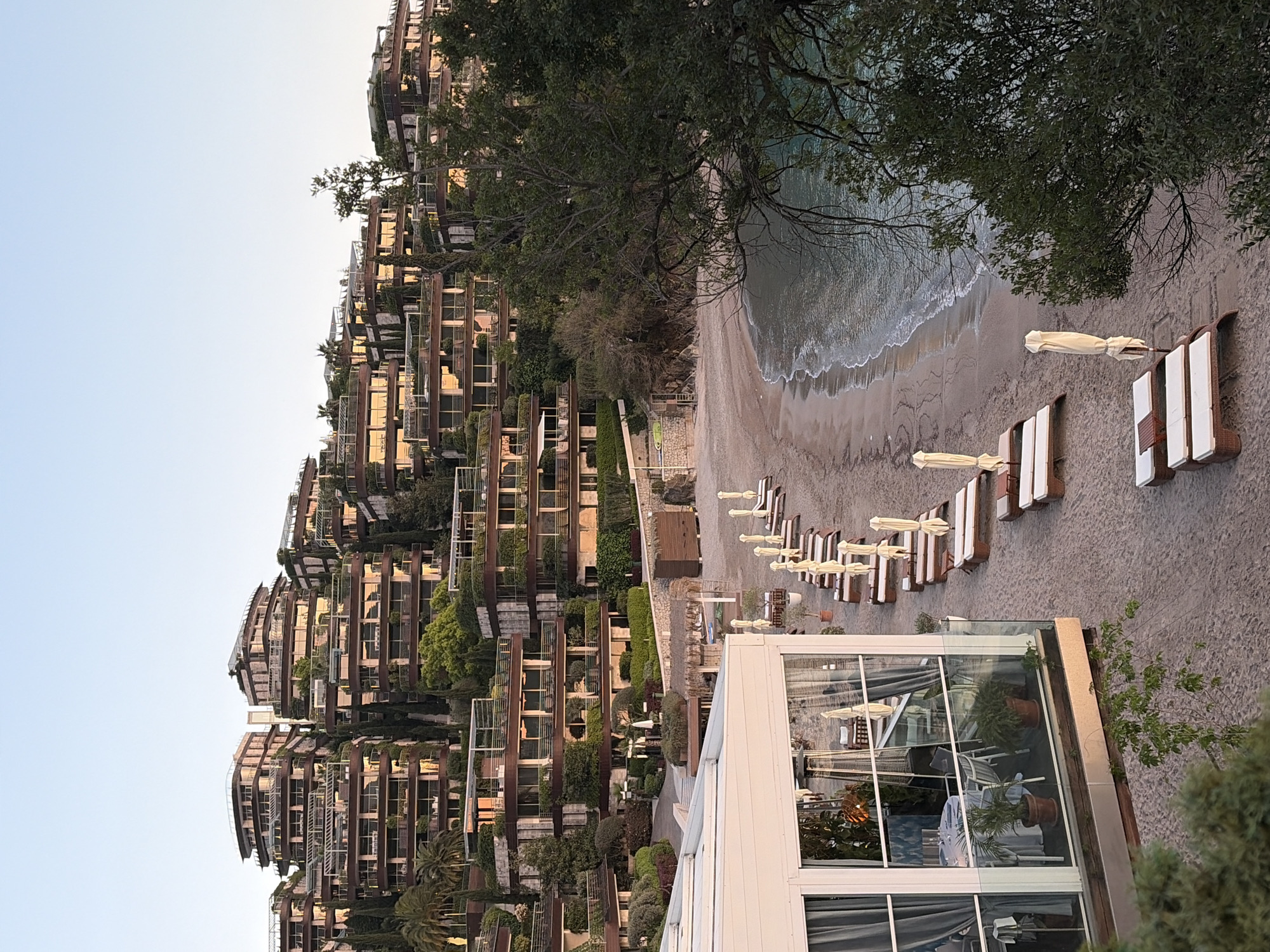 The image shows a resort complex in Budva, Montenegro. The architecture is modern with multiple tiered buildings featuring balconies covered in greenery. A beach is visible with lounge chairs and sun umbrellas arranged along the shoreline. Waves are gently lapping the sand. In the foreground, there is a glass-enclosed structure, possibly a restaurant or lounge area, with plants inside. The sky is clear, indicating a sunny day.