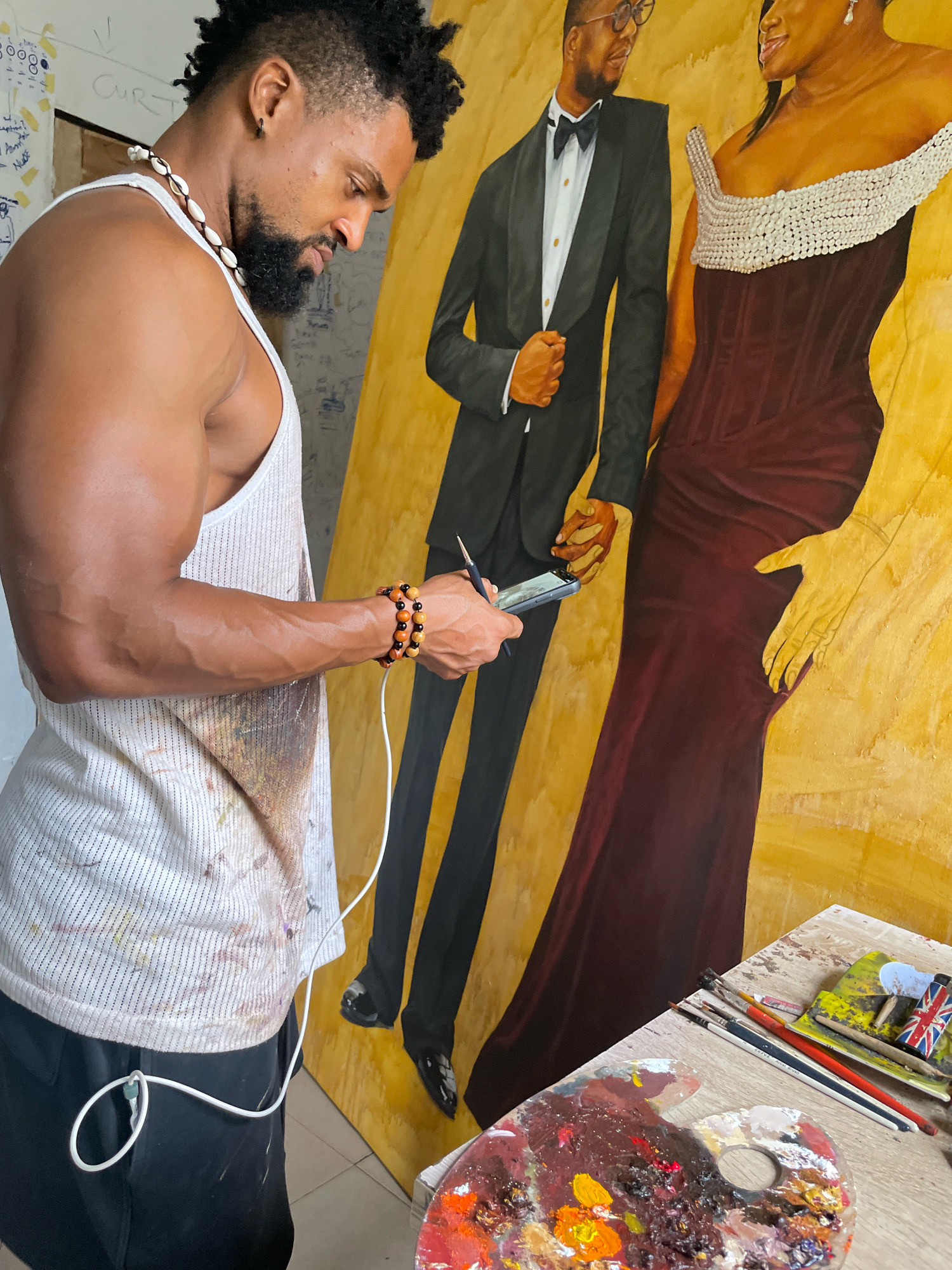 An artist, a Black male with a mohawk haircut, beard, and wearing a white tank top adorned with paint splatters, stands in a studio in Ado, Nigeria. He is in the process of painting a large canvas depicting a Black couple in formal attire. The male figure in the painting wears a tuxedo, and the female figure wears a burgundy dress with a pearl-embellished bodice. The artist holds a paintbrush and a smartphone in his hands, seemingly referencing the phone for his work. In the foreground, a wooden palette filled with colorful paints and various brushes are laid out on a table. The studio environment appears cluttered, with notes and drawings visible on the wall behind the artist.