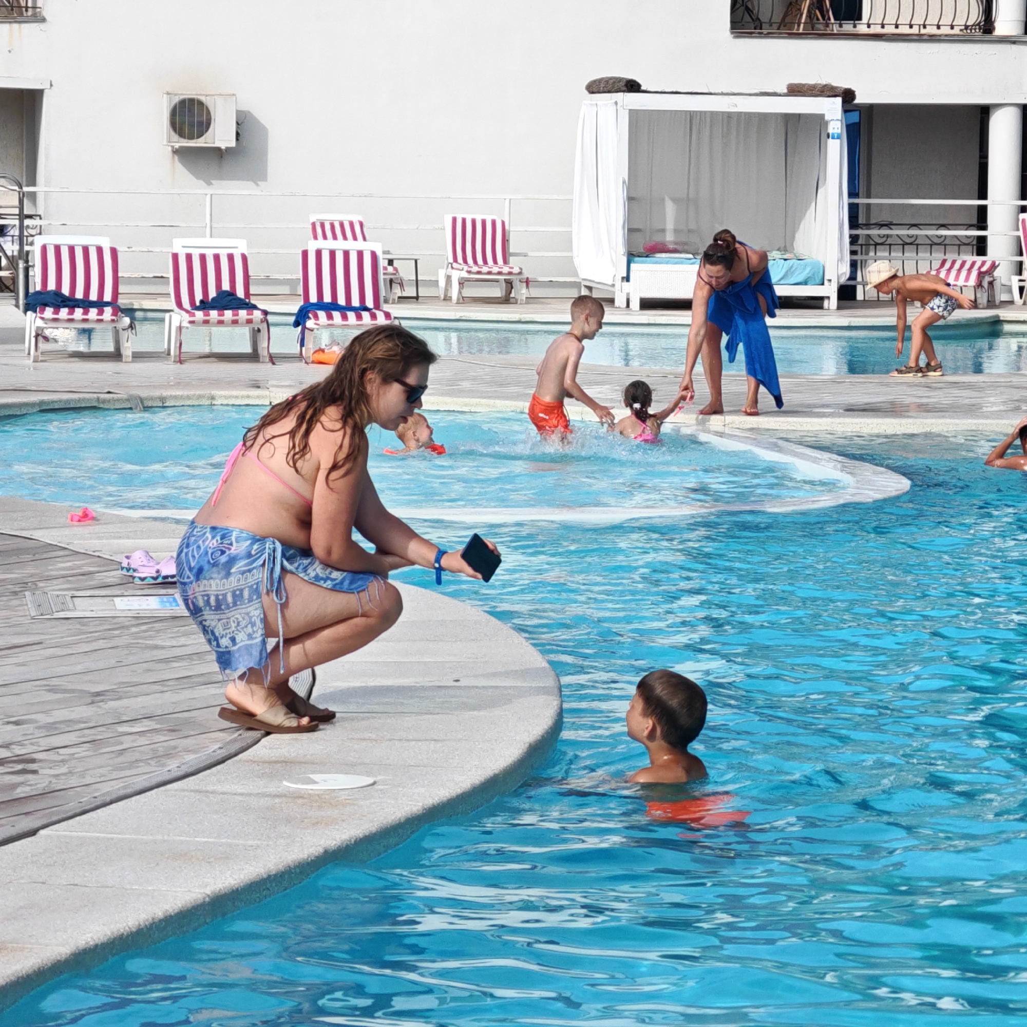 The image depicts a sunny day at a swimming pool. A woman in a pink bikini and a blue patterned sarong is kneeling at the edge of the pool, holding a black smartphone. She appears to be interacting with a young child in the pool. To her right, a young boy in orange swim trunks is in the water, looking up and to the side. In the background, several other people are visible enjoying the pool. There are lounge chairs with striped cushions and a white cabana structure. The pool area appears to be well-maintained, with clear blue water. The location is identified as Мысовское сельское поселение, Ukraine.