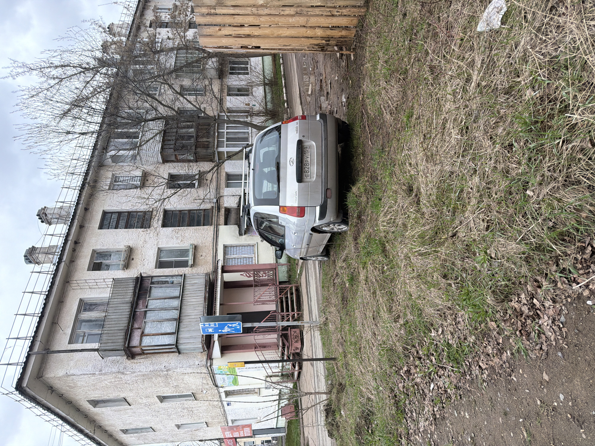 The image depicts a residential scene in the city of Chaikovsky, Russia. A silver Hyundai Getz hatchback is parked on a patch of grassy land next to a multi-story residential building. The building appears to be old, with weathered facades and enclosed balconies. A pedestrian crossing sign is visible near the building's entrance. A wooden fence is erected behind the car, partially obscuring a view of the surroundings. The ground consists of dry grass and dirt. The sky is overcast, suggesting dull weather conditions. The license plate of the car is visible as "E828PY 159".