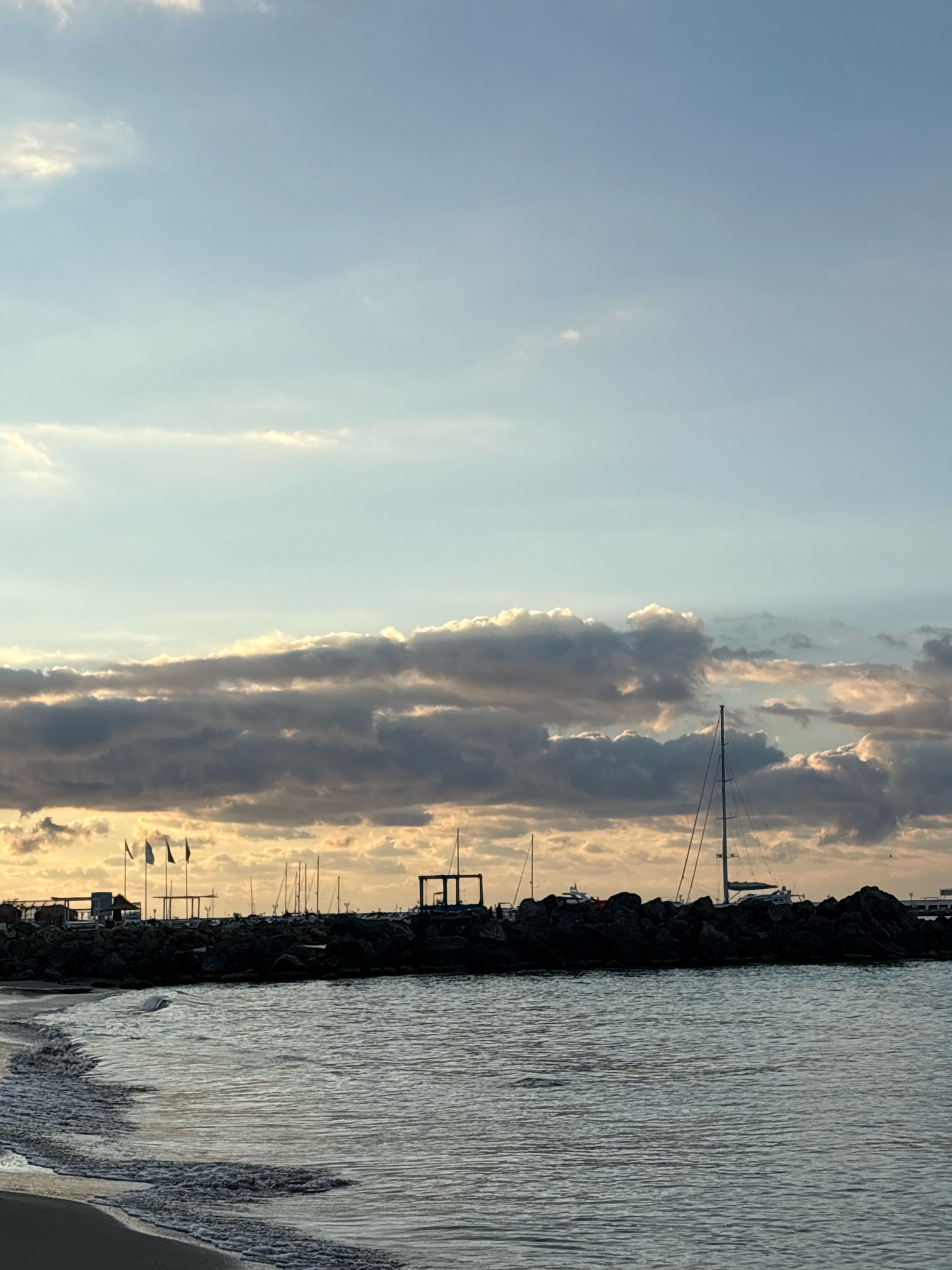 The image shows a view of a harbor in Sveti Vlas, Bulgaria, at sunset. The sky is filled with dramatic clouds tinged with orange and yellow from the setting sun. A stone breakwater, partially covered in shadows, forms a barrier between the water and the harbor. On the breakwater, several boats are docked, including sailboats with tall masts. In the background, there are some buildings and flags. The sea in the foreground is calm, with gentle waves lapping at a sandy beach on the left side of the frame. The overall scene conveys a peaceful and picturesque end to the day.