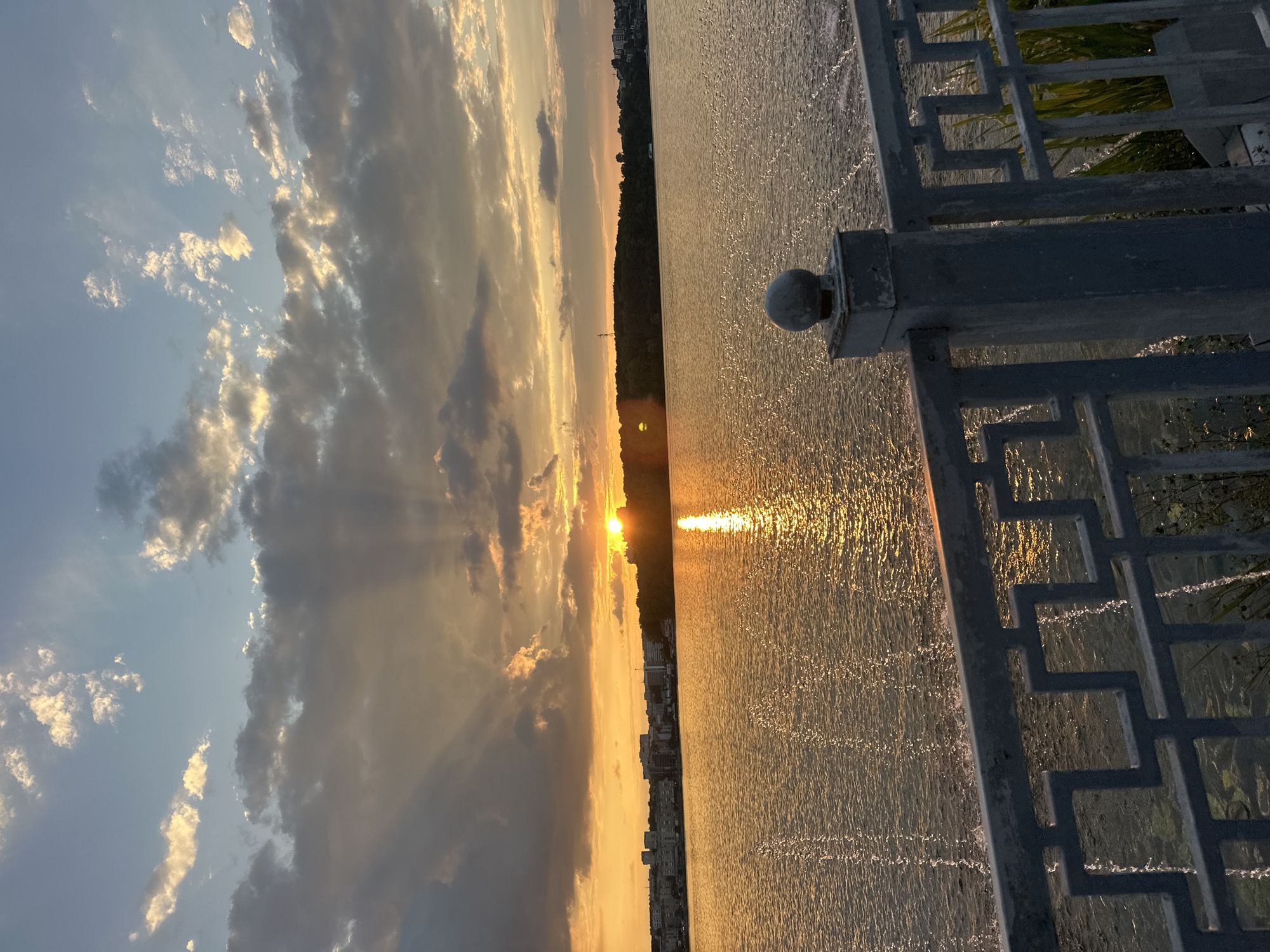 The image depicts a sunset over Ternopil Reservoir in Ternopil, Ukraine. The sun, partially obscured by clouds, casts a warm, golden glow across the water and sky, creating a dramatic effect with visible sunbeams radiating downwards. The reservoir stretches across the frame, its surface rippling and reflecting the sunset's light. On the far bank, a line of buildings, characteristic of a city skyline, is visible, nestled behind a dense line of trees. In the foreground, a metal railing with a decorative geometric pattern runs along the edge of what appears to be a promenade or embankment. The railing is weathered, and a supporting post with a rounded cap is visible on the right side of the frame. The overall scene is one of tranquil natural beauty enhanced by the urban landscape.