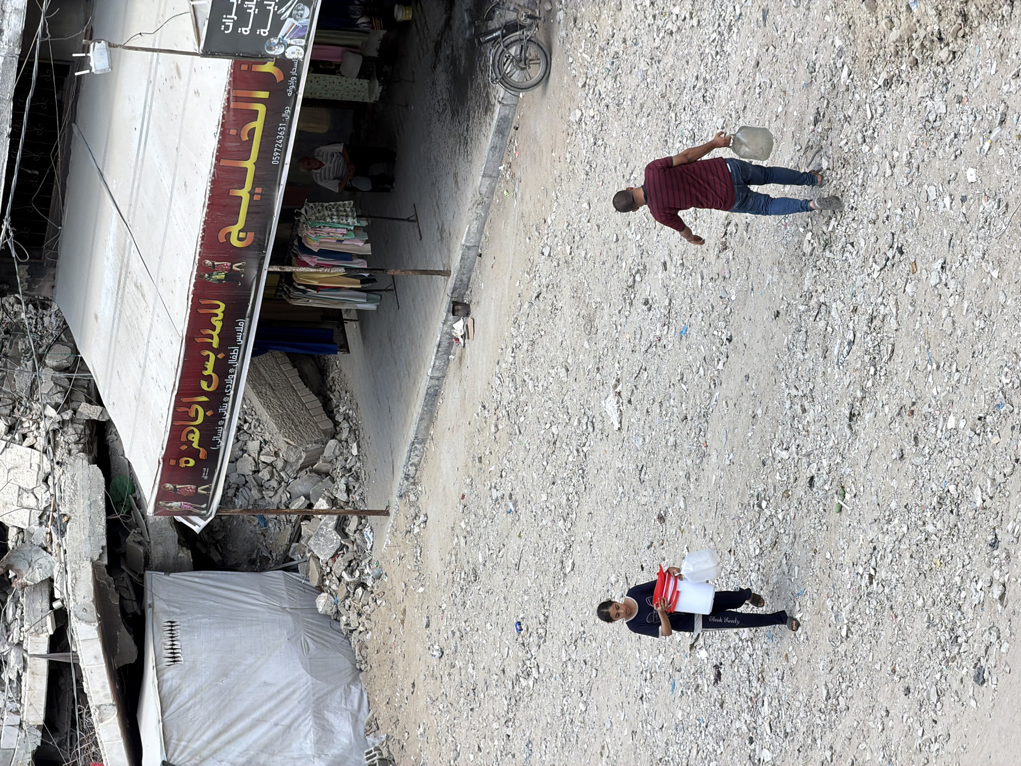 In the city of Gaza, Palestinian Territories, a scene of destruction and resilience unfolds. Buildings lie in ruins, their concrete shells and twisted rebar a stark testament to conflict. Amidst the debris, a small clothing shop, "Al Khaleej Al Gawhara," displays its wares under a faded awning. A man sits in front of the shop, his gaze distant. Nearby, a young girl carries two buckets and a plastic bag, her expression determined as she navigates the rubble-strewn ground. A man in a striped shirt walks away from the camera, also carrying a large plastic jug. A bicycle leans against a curb, a silent witness to the ongoing struggle for normalcy in this war-torn landscape.