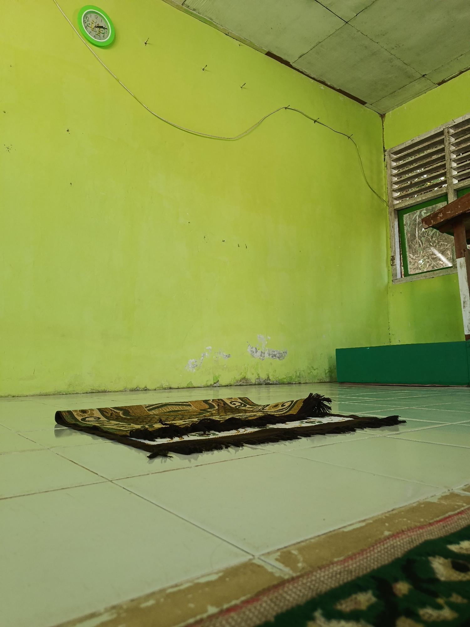 The image depicts an indoor scene, likely a room in Mansalean, Indonesia. The walls are painted a bright lime green, with some areas showing signs of wear and tear. A round, green-framed clock is mounted on the left wall, showing a time between 11:50 and 11:55. A thin electrical wire is strung across the wall, secured by several small clips. On the right wall, there is a louvered window with a wooden frame, through which trees and foliage are visible. A dark wooden table is partially visible in front of the window. On the floor, a prayer mat with a pattern of dark brown, gold, and green is folded and placed on a tiled surface. Another rug, partially visible at the bottom of the frame, appears to be a continuation of the flooring or another mat. A green bench-like object is situated against the wall in the background, to the right of the prayer mat. The lighting suggests it is daytime. There are no people visible in the image.