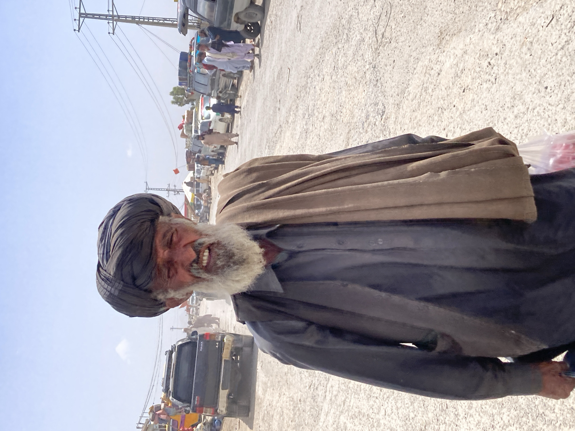 The image captures an outdoor, bright daylight scene in what appears to be a busy, unpaved area, likely a market or roadside gathering in Loralai, Pakistan. The central subject is an elderly man, shown from the chest up, smiling widely towards the viewer. He has a long, thick white beard and is wearing a dark turban, a dark long-sleeved shirt, and a light brown shawl draped over his shoulders. His joyful expression is prominent.
In the background, numerous vehicles are visible, primarily SUVs or 4x4s, with some carrying luggage on their roofs. One specific vehicle in the mid-ground clearly shows "HILUX" and "TOYOTA" branding along with "4x4" on its tailgate. Several other people, mostly men in traditional attire (shalwar kameez), are seen standing or moving around these vehicles, indicating a bustling atmosphere.
The ground is dusty and light-colored, characteristic of an arid or semi-arid region. Electrical utility poles and power lines crisscross the sky in the upper left portion of the image. The sky is clear and bright, suggesting sunny and possibly hot weather. The overall setting depicts a vibrant, everyday scene in a local community. A translucent plastic bag containing red items is partially visible near the man's right arm.