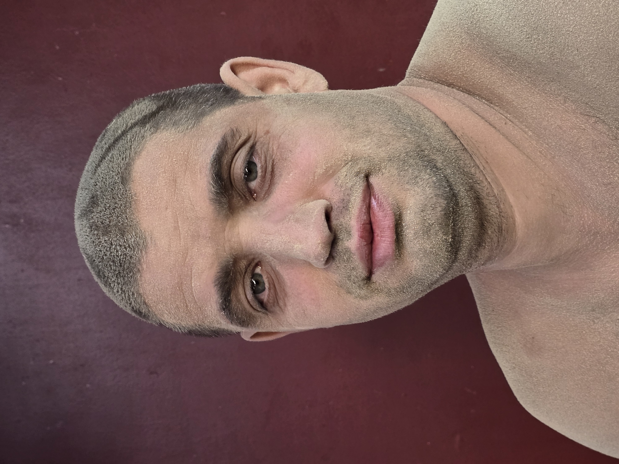 A head and shoulders portrait of a middle-aged man, his skin and short hair lightly dusted with a pale, dry powder. He has a fair complexion, dark eyebrows, and a faint stubble. His eyes are light green, and he is looking directly at the camera with a neutral expression. His lips are slightly parted. The background is a dark, textured red wall. The man appears to be outdoors, possibly in an urban environment, though no specific landmarks of Nova Kakhovka, Ukraine, are visible. The image is sharp and well-lit, focusing on the man's face and upper torso.