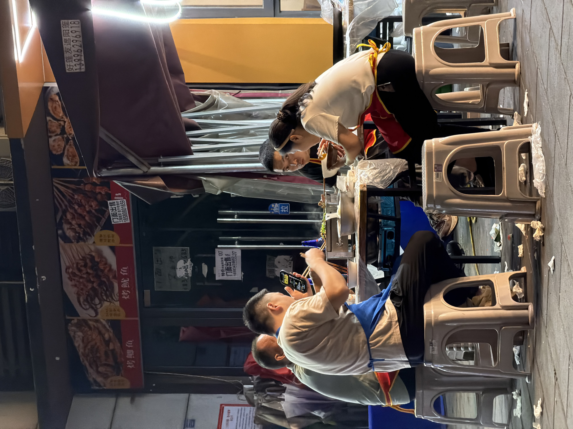 The image captures a bustling street food scene at night, possibly in Chongqing High-tech Zone, China. Several people are seen dining at a food stall. A woman wearing a white shirt, black pants, and a red and yellow apron is focused on preparing or serving food. A man is seated, holding a smartphone in one hand and chopsticks in the other, possibly capturing the meal. The food stall is decorated with images of grilled fish and squid, along with promotional signs. The scene is framed by utilitarian plastic chairs, scattered tissues, and advertisements for businesses like "Haopengyou Sunshade Awning." The lighting suggests a lively atmosphere.