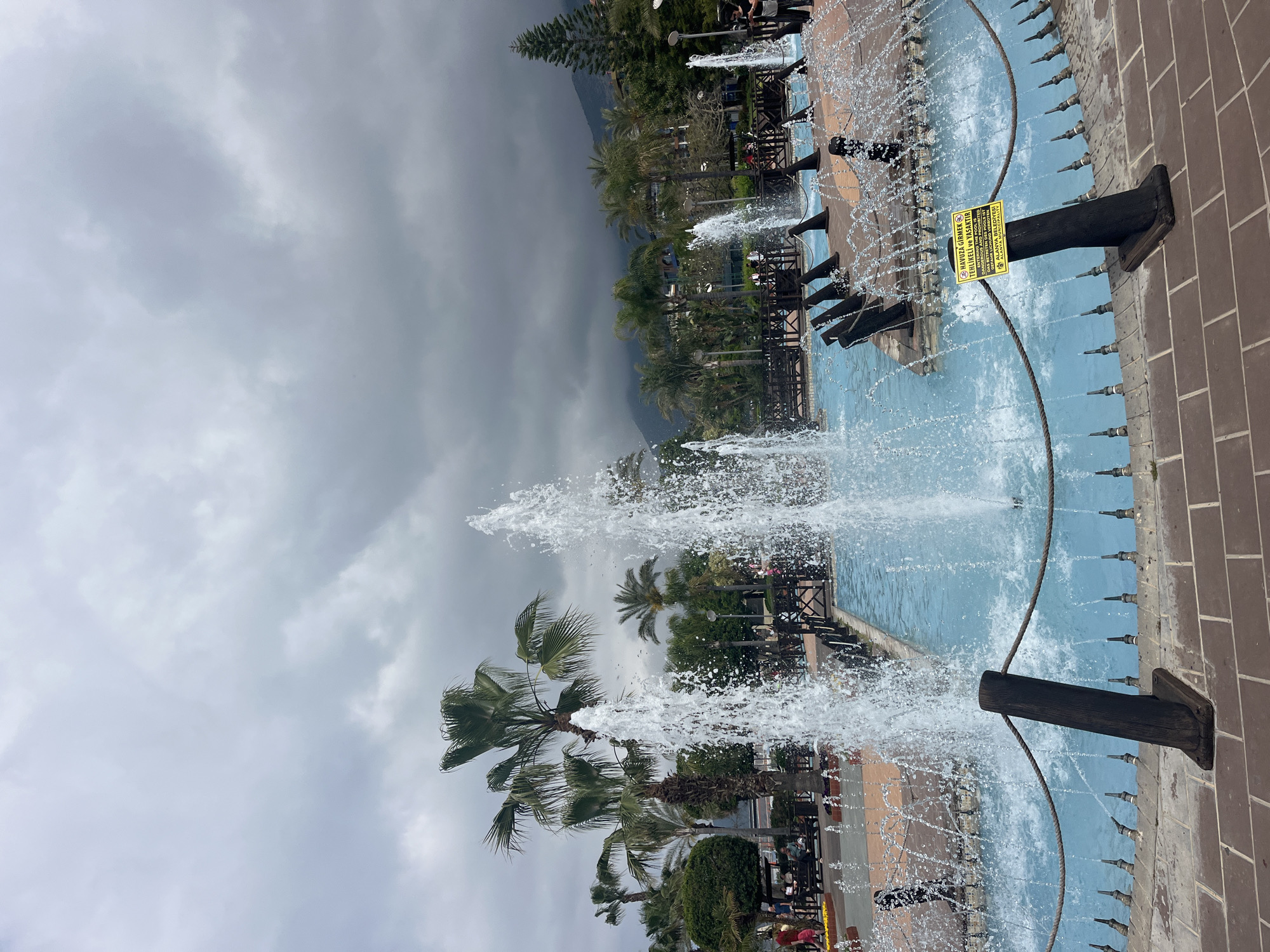The image captures a vibrant outdoor fountain under an overcast sky, likely in Alanya, Turkey. In the foreground, a large, rectangular fountain vigorously sprays water upwards and outwards from numerous nozzles. The fountain pool features light blue tiling and is bordered by a reddish-brown brick pathway. Several decorative elements, resembling dark wooden cannons or spouts, extend from the brick edge into the fountain, contributing to the water display. A thick rope-like barrier also runs along the edge of the fountain.
A prominent yellow warning sign is attached to one of the wooden cannon-like structures. The text on the sign, in Turkish, English, and Russian, states: "HAVUZA GİRMEK TEHLİKELİ VE YASAKTIR. SWIMMING IN THE FOUNTAIN IS DANGEROUS AND PROHIBITED. ОТКРЫТАЯ ВХОД В БАССЕЙН ОПАСНО И ЗАПРЕЩЕНО." It also bears the inscription "ALANYA BELEDİYESİ" (Alanya Municipality).
Beyond the fountain, lush green palm trees and other dense foliage line a paved area, suggesting a park or public garden setting. Faint figures of people can be seen in the background, some seated, others standing. In the distance, dark, imposing mountains rise against a dramatic, heavily clouded sky, suggesting either an impending storm or very heavy overcast conditions during the daytime. The scene combines natural elements with man-made landscaping and an active water feature, conveying a sense of a public recreational space.