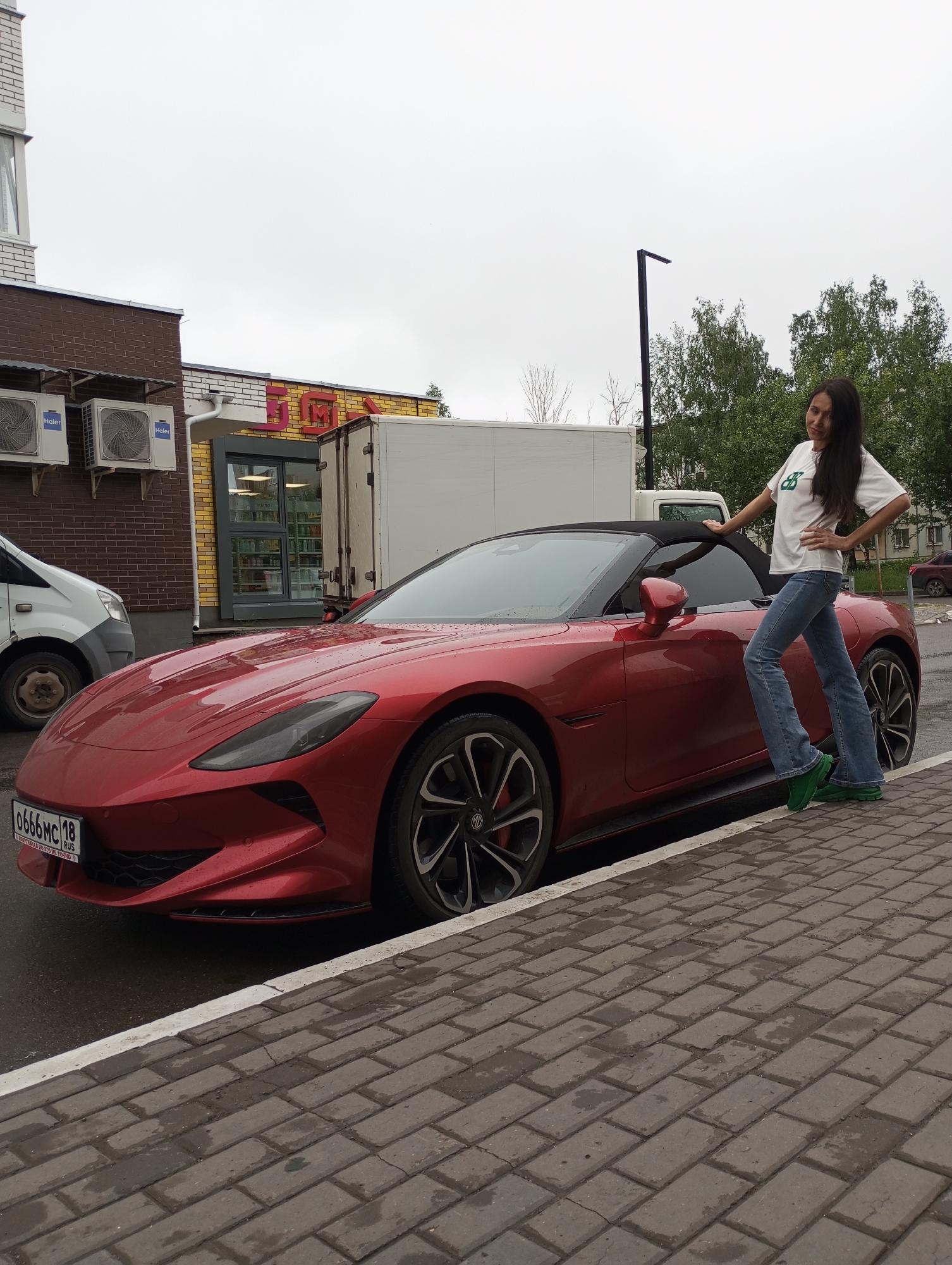 The image shows a woman standing next to a red MG Cyberster convertible in a city. The license plate reads "0666MC 18 RUS," indicating that the location is likely Izhevsk, Russia, part of the Udmurt Republic (code 18). The car is parked on the side of the road, next to a brick sidewalk. The woman is wearing a white t-shirt with a green design, blue jeans, and green shoes. Behind the car and the woman, there is a building with what appears to be a storefront (marked "MAGNIT" in Russian) and a white delivery van. The sky is overcast, suggesting it is a cloudy day. There are trees in the background as well.