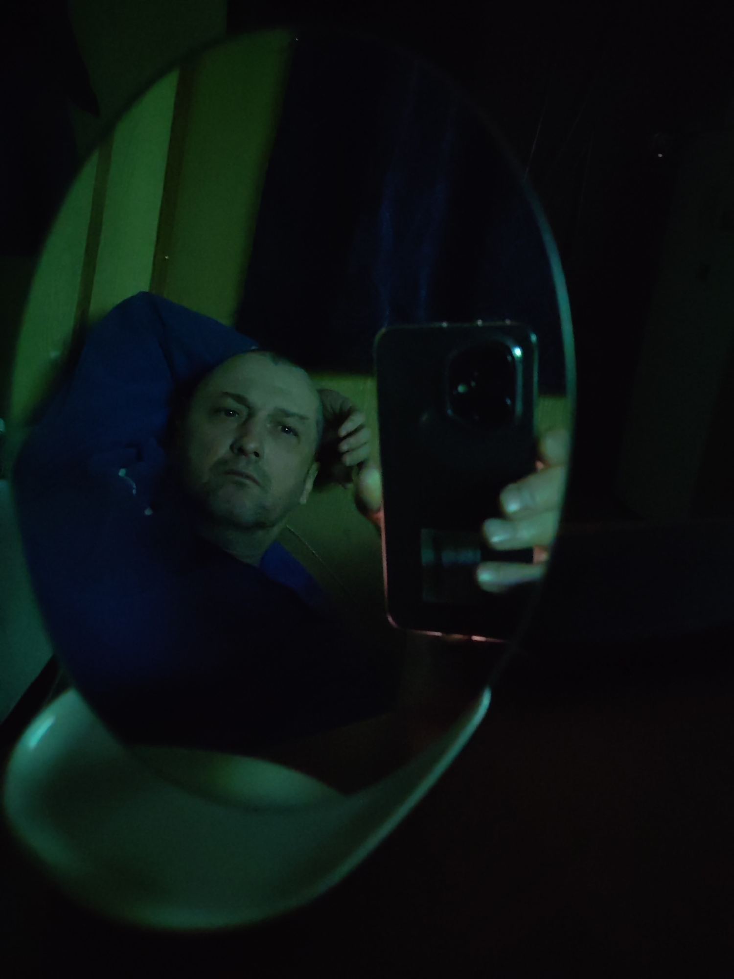 An image captures a mirror selfie of a man, reflecting his upper body and the smartphone he uses to take the picture. The man, who appears to be middle-aged with short hair, is wearing a dark blue long-sleeved shirt. He is looking directly into the camera, which is reflected in the oval mirror. His left arm is bent, with his hand resting near his head, suggesting a relaxed posture. The smartphone he holds is dark-colored, possibly black, with visible camera lenses on its back.
The scene is dimly lit, with a prominent green hue originating from the left side and background, illuminating what appears to be a wall or paneling behind the man. This green light creates a moody and atmospheric setting. The overall background outside the mirror's reflection is dark. A light-colored, curved object, possibly part of a chair or table, is visible at the bottom of the mirror's reflection. The time of day appears to be evening or night, given the low light conditions. The man's expression is neutral or contemplative. No visible text is present in the image.