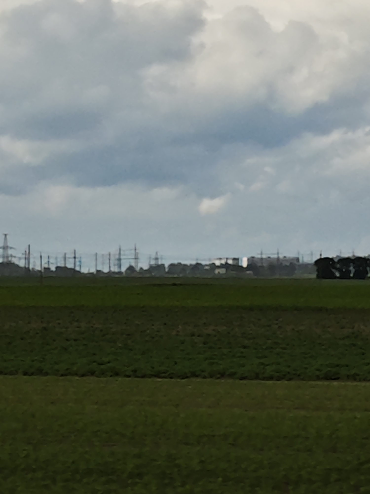 The image depicts a landscape scene with a focus on power lines stretching across the horizon. The foreground is dominated by two distinct bands of green fields, suggesting agricultural land. The upper band appears to be a darker, more cultivated field, while the lower band is a lighter, possibly fallow or less dense field.
In the middle ground, a line of numerous tall power line pylons, interconnected by wires, forms a prominent feature against the sky. These structures are silhouetted against a hazy, overcast sky. Interspersed with the pylons are hints of buildings and trees, creating a sense of a distant settlement or industrial area. The overall impression is of a somewhat industrial and rural environment.
The sky is filled with heavy, grey clouds, indicating a cloudy or possibly overcast day. The light is diffused, suggesting it is not a bright, sunny day, and likely late afternoon or early morning, or simply a heavily overcast day. There are no people visible in the image, and the focus is on the landscape and the man-made structures within it. The scene conveys a sense of vastness and the presence of infrastructure in a relatively open landscape. The location context provided is Красненскі сельскі Савет, Belarus, which places this scene within a specific rural area in Belarus. There is no visible text in the image.