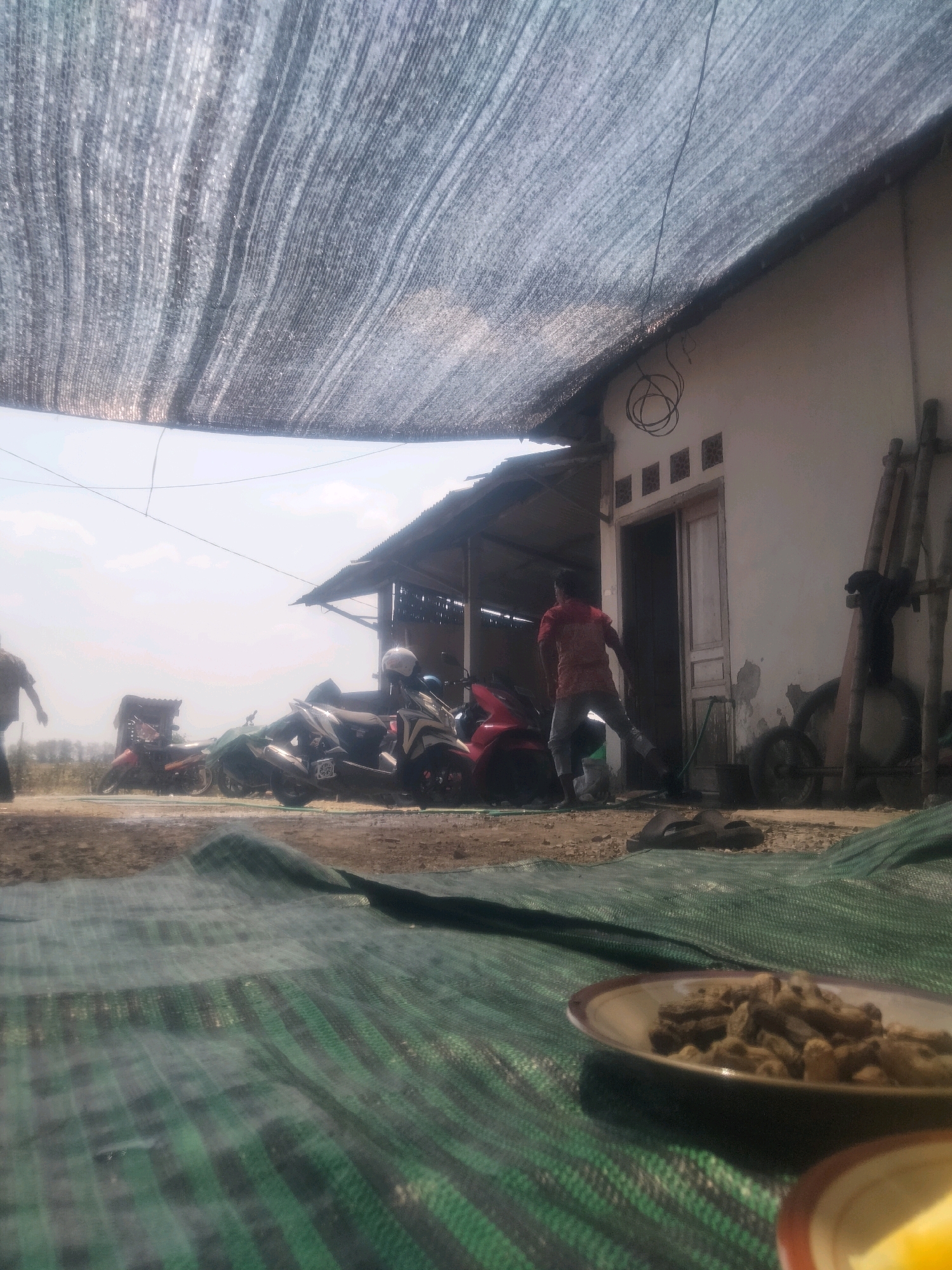 The image captures an outdoor scene in Soko, Indonesia, likely a rural area or the outskirts of a town, characterized by a simple dwelling and a yard. A dark, woven shade cloth stretches overhead, casting dappled shadows. In the mid-ground, several motorcycles are parked, some covered with green tarps. To the right, a light-colored building with an open doorway and a small, open-air shelter with a corrugated metal roof are visible. A man in a red shirt and gray pants is near the building entrance, holding a green hose. To the left, another person, partially visible and out of focus, walks away from the camera. In the foreground, a green, patterned ground cover is laid out, with a plate of what appears to be dried food items in the lower right corner. The environment appears dusty and dry, suggesting a warm climate. The overall impression is one of a casual, everyday setting.