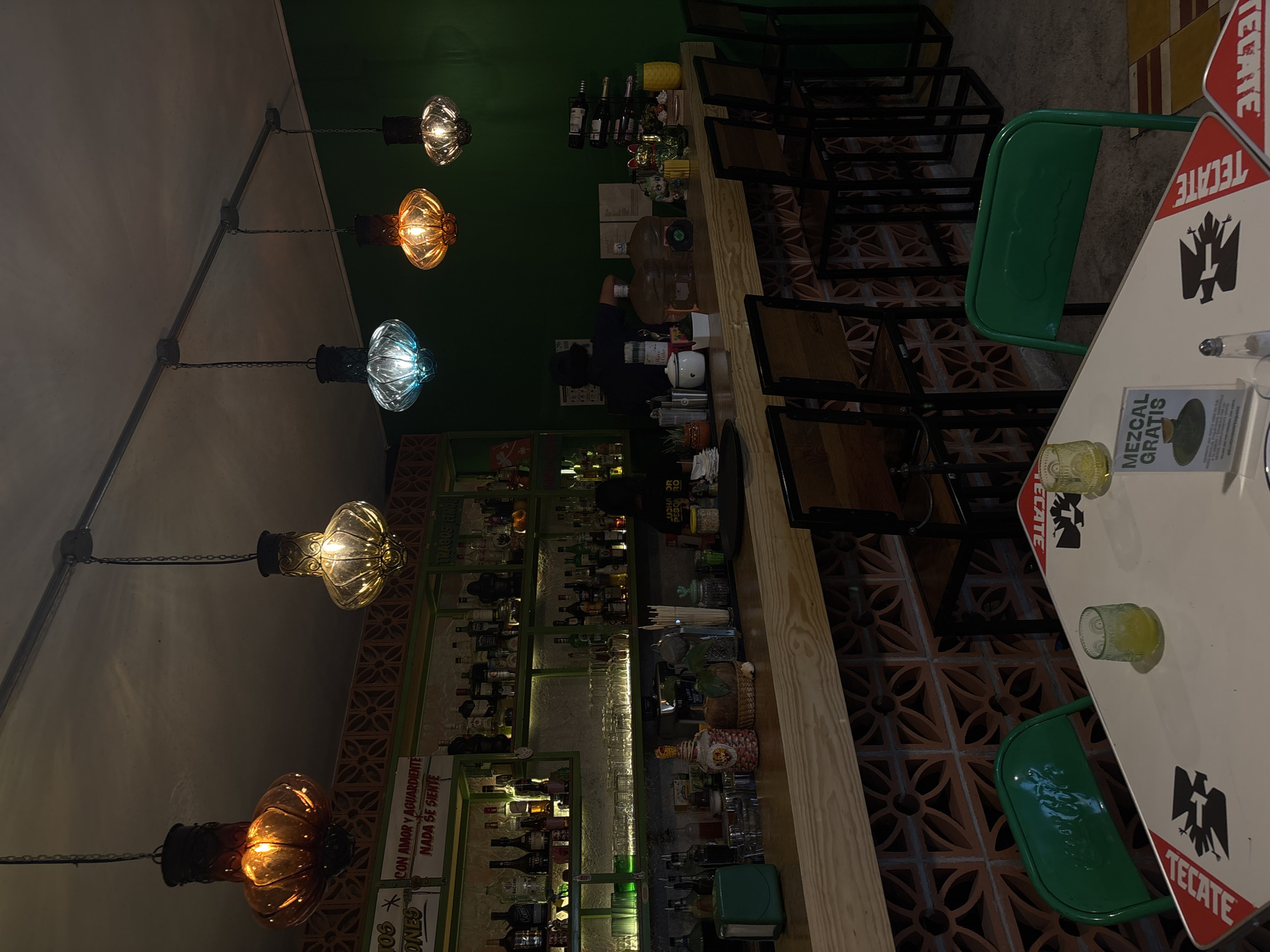 This image depicts the interior of a bar or cantina in San Andrés Cholula, Mexico, during what appears to be evening hours. The scene is dominated by a long, light-colored wooden bar counter that runs horizontally across the mid-ground.
**Environment:**
The walls are painted a dark green color. Behind the bar, a series of open shelves with green frames are visible, illuminated from below by strip lighting, showcasing numerous bottles of liquor and glassware. The lower portion of the bar front features a decorative pattern of terracotta-colored bricks. The ceiling is dark, from which five ornate, glass pendant lamps hang at varying heights; two are amber, one is blue, and two appear clear or amber-toned with internal light sources. The floor in the visible areas is a light grey concrete or similar material, with some patterned tiles on the right.
**Subjects and Actions:**
One individual, presumably a bartender, is positioned behind the bar on the right side of the frame, facing away from the camera. The person is wearing a dark shirt and is seen reaching towards or interacting with items on the bar counter. No other patrons are visibly present or interacting.
**Objects:**
* **Bar Counter:** Cluttered with various bar tools, mixing glasses, several large plastic water carafes, a white ceramic container, a small plant, and a napkin dispenser. Bottles of alcohol are densely arranged on the shelves behind the bar, including brands like Bacardí, Malibu, and what appear to be various tequilas and mezcals. A sign reading "CON AMOR Y AGUARDIENTE NADA SE SIENTE" is visible above the left section of the back bar, and "TRAGOS FINOS" is above the right section.
* **Seating:** A row of at least five empty bar stools with dark metal frames and wooden seats are lined along the bar. In the foreground, two small, rectangular tables are visible. These tables are white with red borders and feature "TECATE" branding and an eagle logo. Two green metal chairs are placed at these tables.
* **Table Objects:** On the table closer to the viewer, two small, textured clear glasses are present; one contains a yellow liquid. A small metal salt shaker and a vertical sign that reads "MEZCAL GRATIS" (Free Mezcal) are also on this table.