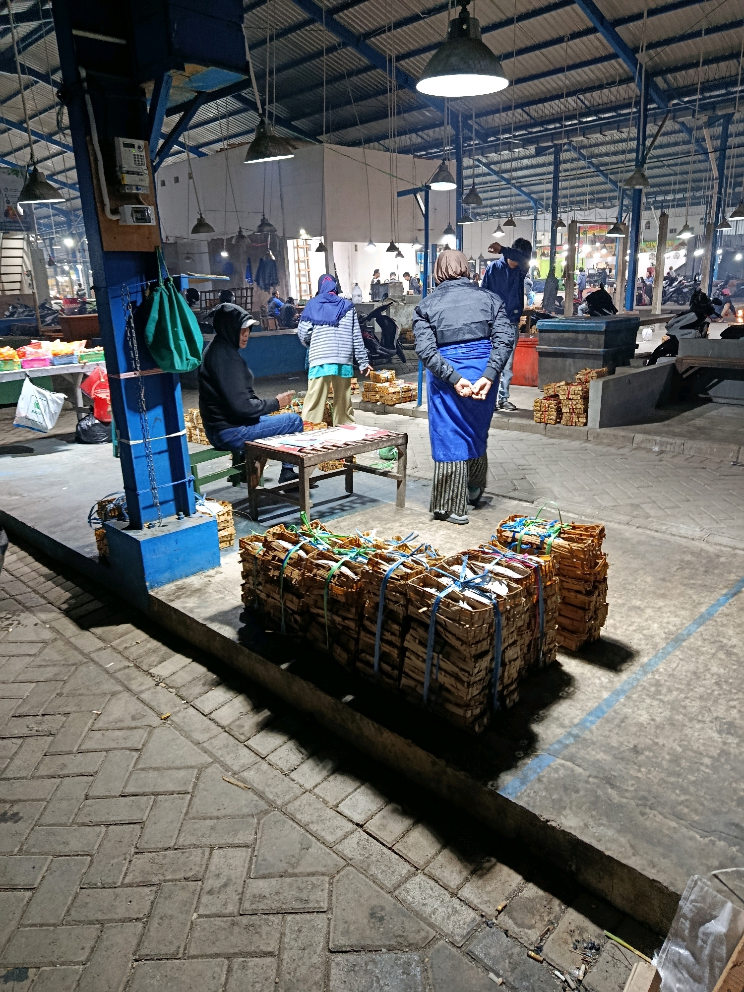 The image depicts a dimly lit indoor market, likely in Malang, Indonesia. The ceiling is a metal structure with exposed beams and industrial lighting fixtures. In the foreground, several stacks of tightly bound goods, possibly firewood or similar items, are arranged on the concrete floor. These stacks are secured with blue and green ropes. To the left, a person wearing a black hooded jacket and jeans sits on a green stool at a wooden table, looking towards the right. A green bag hangs from a blue metal post near them. Several more stacks of bound items are visible at the base of this post.
In the mid-ground, two individuals are visible. One person, wearing a dark jacket and a blue skirt or wrap, stands with their back to the camera, hands clasped behind their back. Another person, wearing a blue garment and a headscarf, is walking away. Further back, more people are scattered throughout the market, some interacting near stalls and displays, and a few motorbikes are parked. The floor is a combination of patterned concrete paving blocks and smooth concrete. The overall atmosphere suggests a bustling market environment during evening hours.
