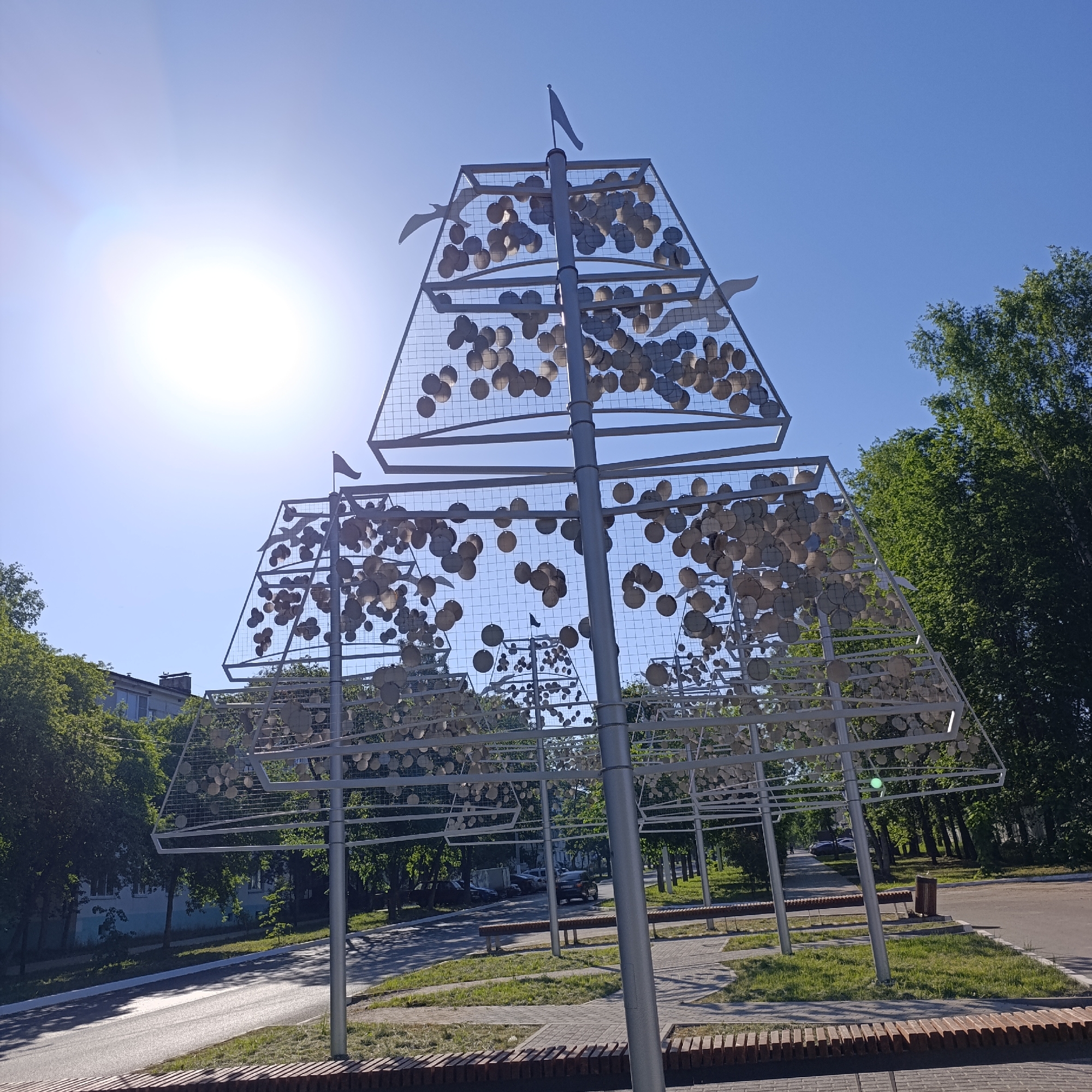 The image captures an outdoor scene in the city of Chaikovsky, Russia, under a bright, clear sky with the sun shining. The central subject is a modern art installation, a metal structure resembling a stylized Christmas tree or tiered pyramid. The structure consists of metal frames arranged in three triangular layers, each filled with a grid of small, round, metallic discs or plates that appear to be hanging freely. Several metal sculptures of seagulls are attached to the frame. A small metal flag is on the top of the structure.
The installation is located at a park or public space adjacent to a road. The road is asphalt and appears to be well-maintained. A low brick wall serves as a border between the road and the park area. Green grass and various trees surround the installation, giving the scene a natural, outdoor feel. A building can be seen in the background.