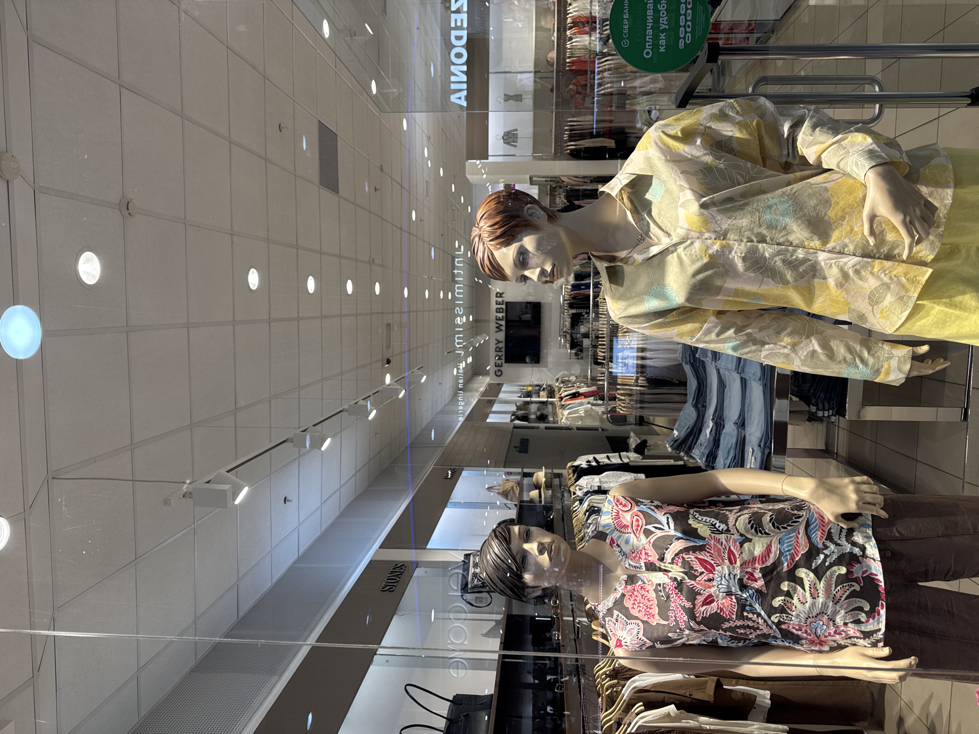 The image captures the interior of a clothing store, specifically focusing on the display window. Two mannequins are prominently featured, dressed in contemporary attire. The upper mannequin wears a yellow floral-patterned jacket and yellow pants, while the lower one sports a brown top with a vibrant floral design and brown pants. Garments hang on racks behind the mannequins, revealing the store's inventory, which includes jeans and other clothing items. The shop's name, "GERRY WEBER," is visible in reverse due to the reflection on the store window, along with the names "SERDONIA" and "STONES." The background reveals a long aisle lined with clothing racks, illuminated by recessed ceiling lights. A sign with green text and a logo in Russian is also visible, indicating that the store is located in a Russian-speaking region. The city and country are Unknown.