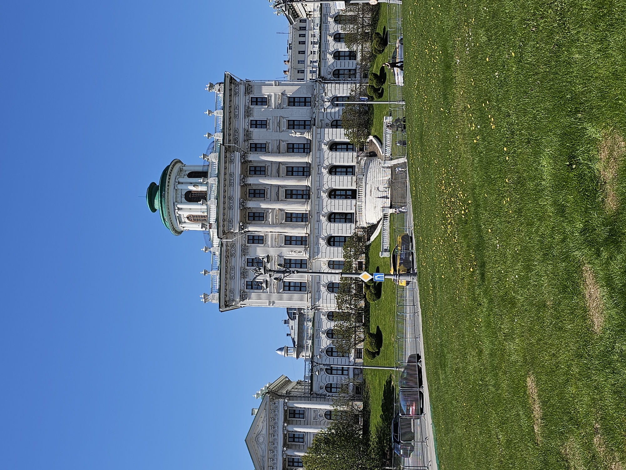The image shows the Senate Palace in the Moscow Kremlin. The building is white with several stories and many windows. It features a green-domed rotunda. The sky is clear and blue. There is a green lawn with some yellow flowers on the right side of the image. A metal fence separates the building and lawn from a road where several cars are parked. A pedestrian walks along the sidewalk.