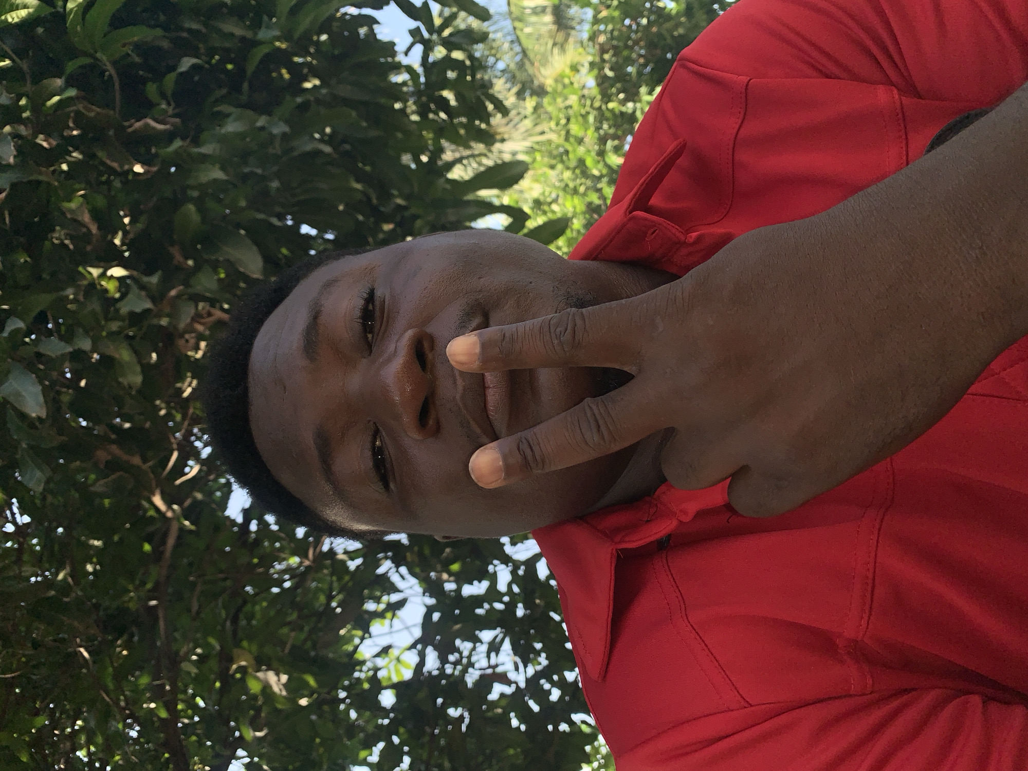 A close-up, medium shot shows a Black man with dark skin and short, black hair posing for a selfie outdoors. He is wearing a red collared shirt with visible stitching. His right hand is raised, with his index and middle fingers extended in a peace sign, positioned near his face. The man is looking towards the camera with a slight smile. Behind him, lush green foliage and dappled sunlight suggest an outdoor setting, possibly a park or garden. The man's location is identified as Akwanga, Nigeria.