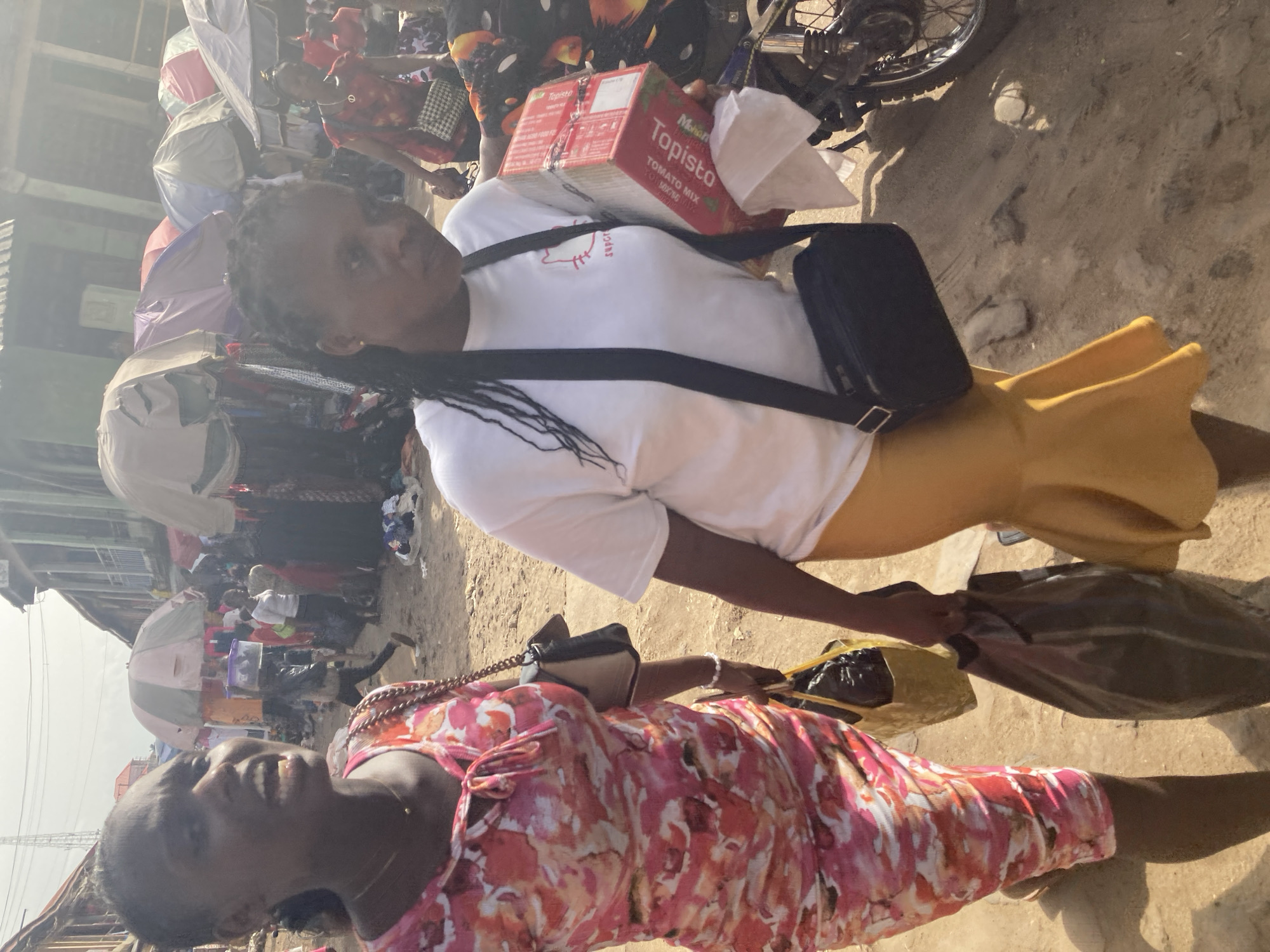 The image depicts a scene in the city of Ura Nta Umuarandu, Nigeria. Two women are visible in the foreground. One woman is wearing a pink and red floral dress and holding a bag. The second woman wears a white t-shirt with a cartoon graphic, a yellow skirt, and a black crossbody bag. She carries a cardboard box labeled "Topisto Tomato Mix." Behind them is a bustling marketplace with numerous people, umbrellas providing shade, and merchandise. A motorcycle is partially visible in the upper right corner of the frame. The ground is unpaved and appears dusty.