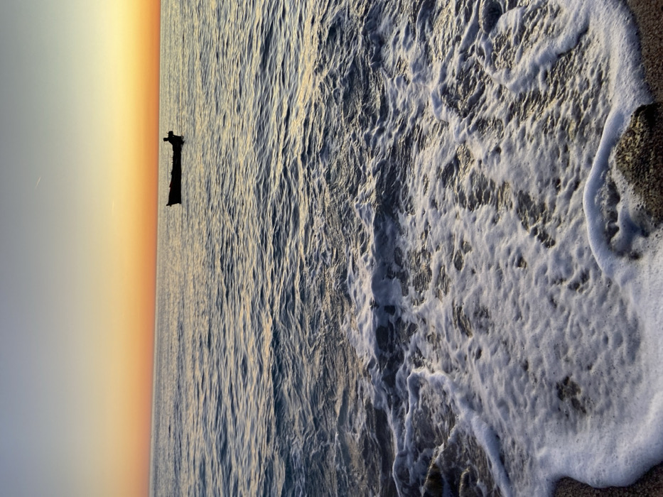 The scene depicts a coastal environment at either sunrise or sunset, in the city of Unknown, Unknown. A low horizon line features a gradient sky, transitioning from bright orange-yellow directly above the horizon to pale blue overhead. The water surface, extending from the mid-ground to the horizon, displays calm, rippling texture with reflections of the sky. Approximately one-third up from the bottom of the image, a dark, small, silhouetted boat is present on the water, containing a single, indistinct human figure, also silhouetted. In the foreground, white seafoam from breaking waves covers a dark, sandy beach interspersed with several dark, irregular rock formations. A faint linear contrail is visible in the upper left quadrant of the sky. No other subjects or structures are discernible.
