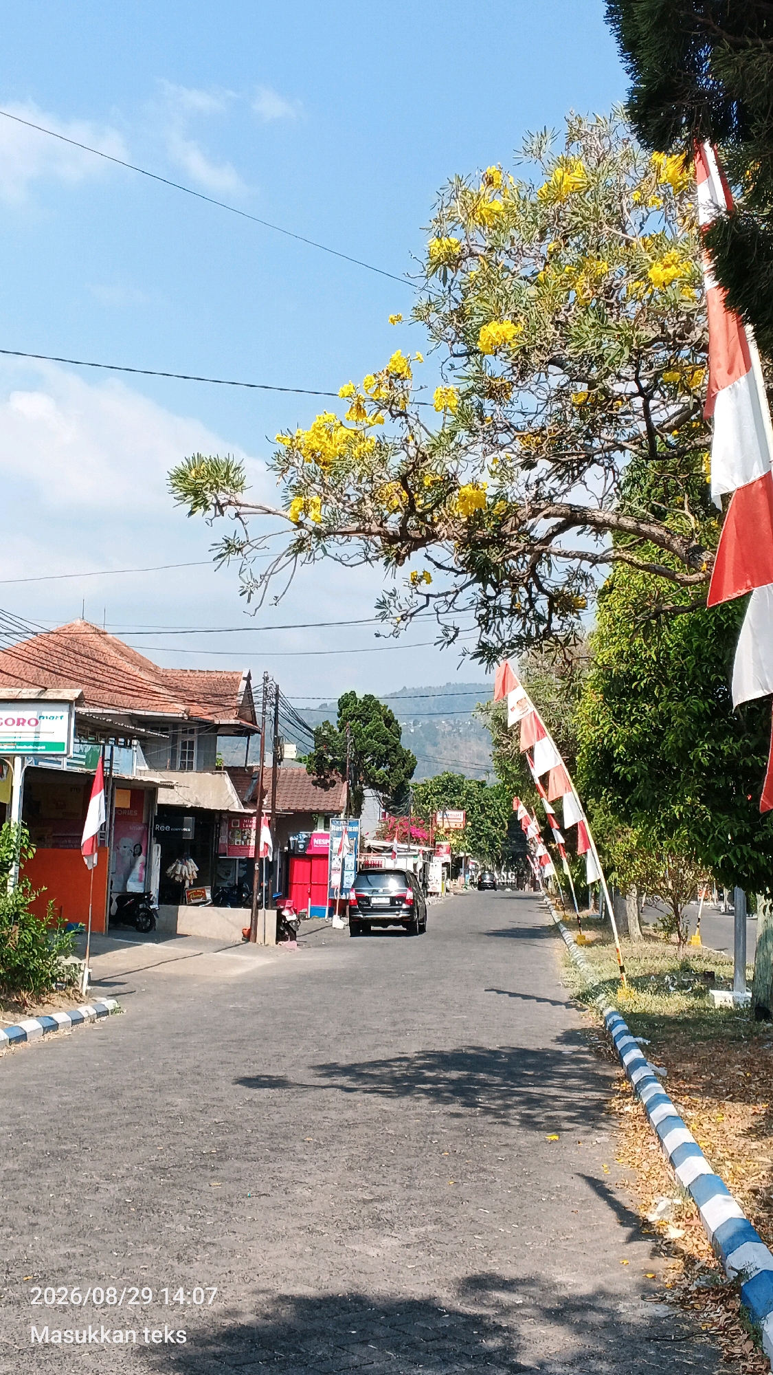 The image depicts a street scene in Sumberporong, Indonesia. A black SUV is driving down a paved road lined with trees and buildings. Red and white flags are hung along the side of the road, suggesting a festive occasion or national celebration. The weather appears to be sunny with a clear blue sky. There are some scattered leaves on the ground, indicating it may be autumn or a time of year with falling leaves. A watermark at the bottom left corner shows "2026/08/29 14:07" and "Masukkan teks" which translates to "Enter text".