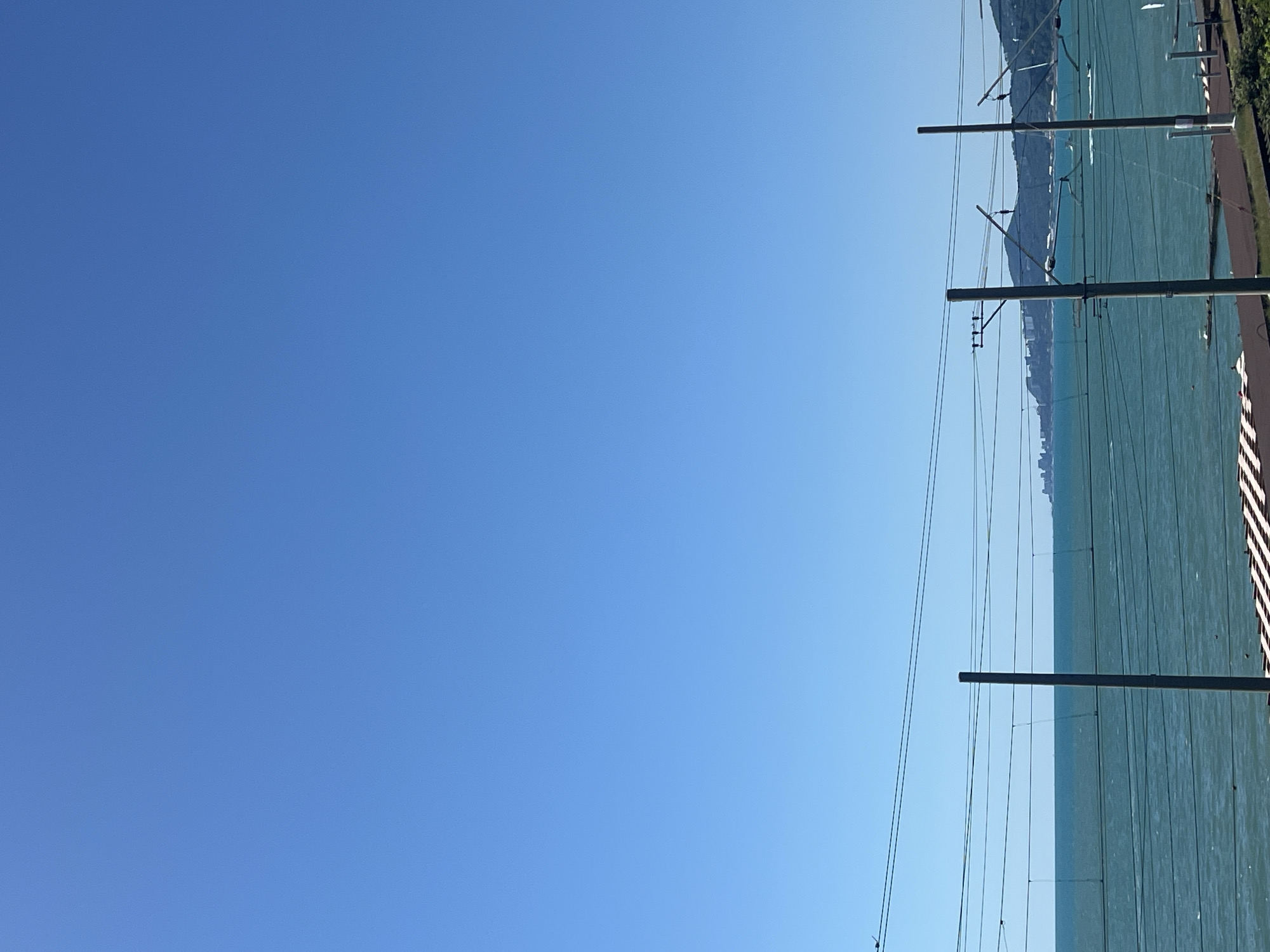 This image captures a bright, sunny day with a clear blue sky over the Black Sea. In the foreground, there are visible train tracks and overhead power lines supported by tall poles. The sea is a vibrant turquoise, with small whitecaps indicating a light breeze. In the distance, along the coastline, is a densely built urban area with buildings that appear to be part of the city of Sochi, Russia. The shoreline is lined with green, mountainous terrain. The scene suggests a coastal railway line running along the edge of the city. There are no people or vehicles immediately visible in the frame, but the presence of the urban development and the water implies activity.