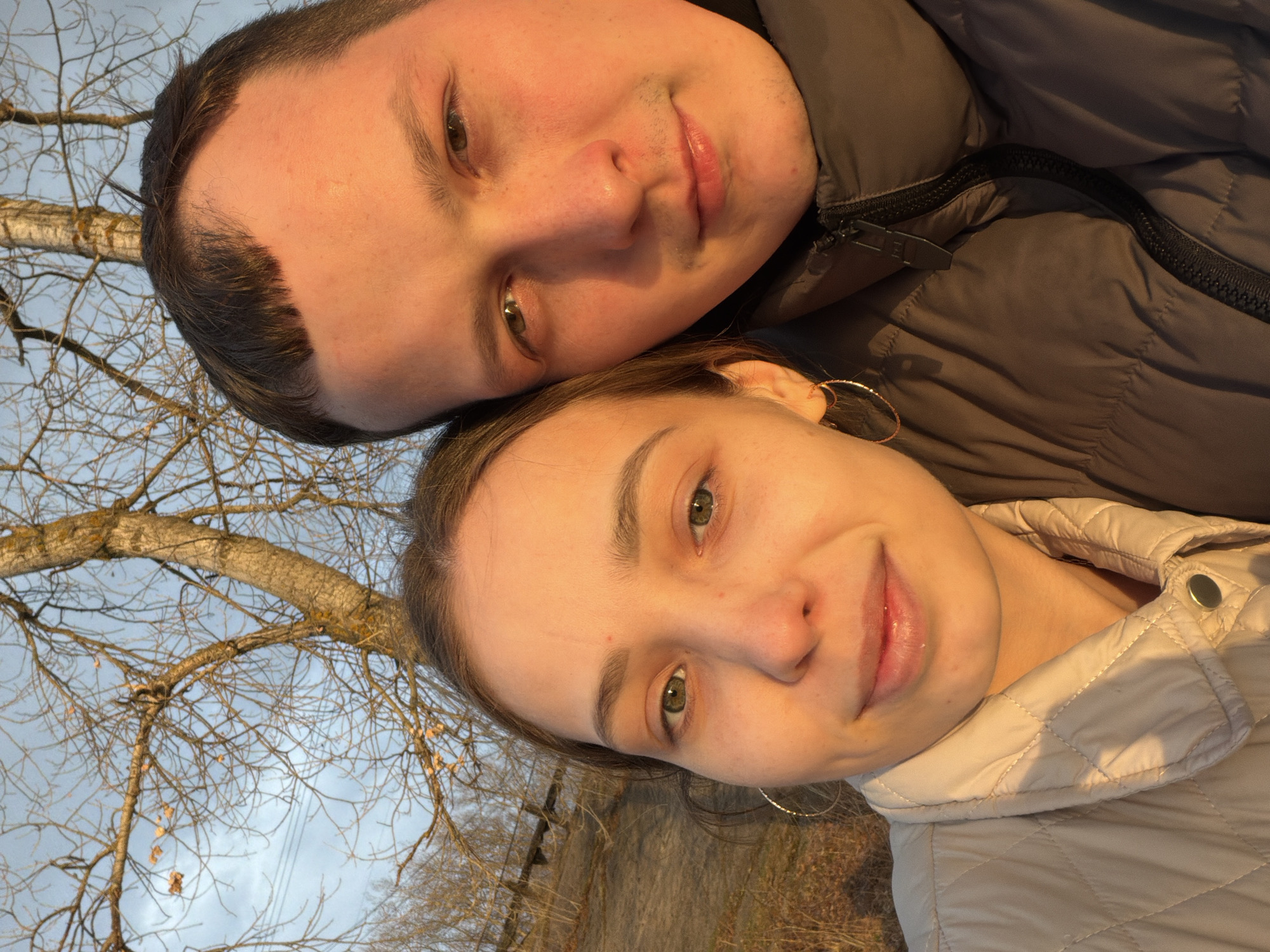 This close-up, upward-angled portrait features two young adults, a male and a female, positioned closely together outdoors. Their heads are tilted, and they are looking directly at the camera with soft, relaxed smiles, conveying a sense of contentment and affection.
The male, positioned on the upper left, has short dark hair and light brown eyes. He is wearing a dark grey or black puffer jacket with a visible zipper. The female, on the lower right, has lighter brown hair and striking green eyes. She wears a light, possibly off-white or beige, quilted jacket with a snap closure, and a delicate hoop earring is visible on her left ear.
The lighting suggests late afternoon or early evening, with a warm, golden glow illuminating their faces. The background consists of a clear or partly cloudy sky, with numerous bare tree branches extending across the frame, indicating an autumn or early winter season. A few dried leaves still cling to the branches. Hints of dry ground or sparse vegetation are visible beneath the female. The setting appears to be a natural or semi-natural outdoor environment, aligning with the location context of Chaikovsky, Russia. There is no visible text.
