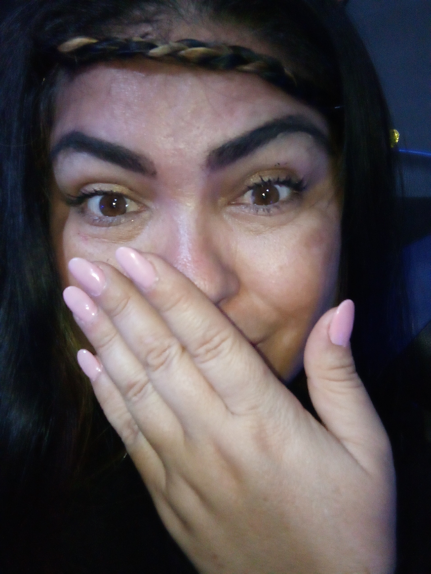 A close-up portrait shows a woman with dark hair and brown eyes. She wears a braided headband, has arched eyebrows, and light pink almond-shaped nails. She covers her mouth with her hand. The background is dark, giving the impression of an indoor setting. No information in the image indicates a specific location. The city of Juan Griego, Venezuela, cannot be ascertained.