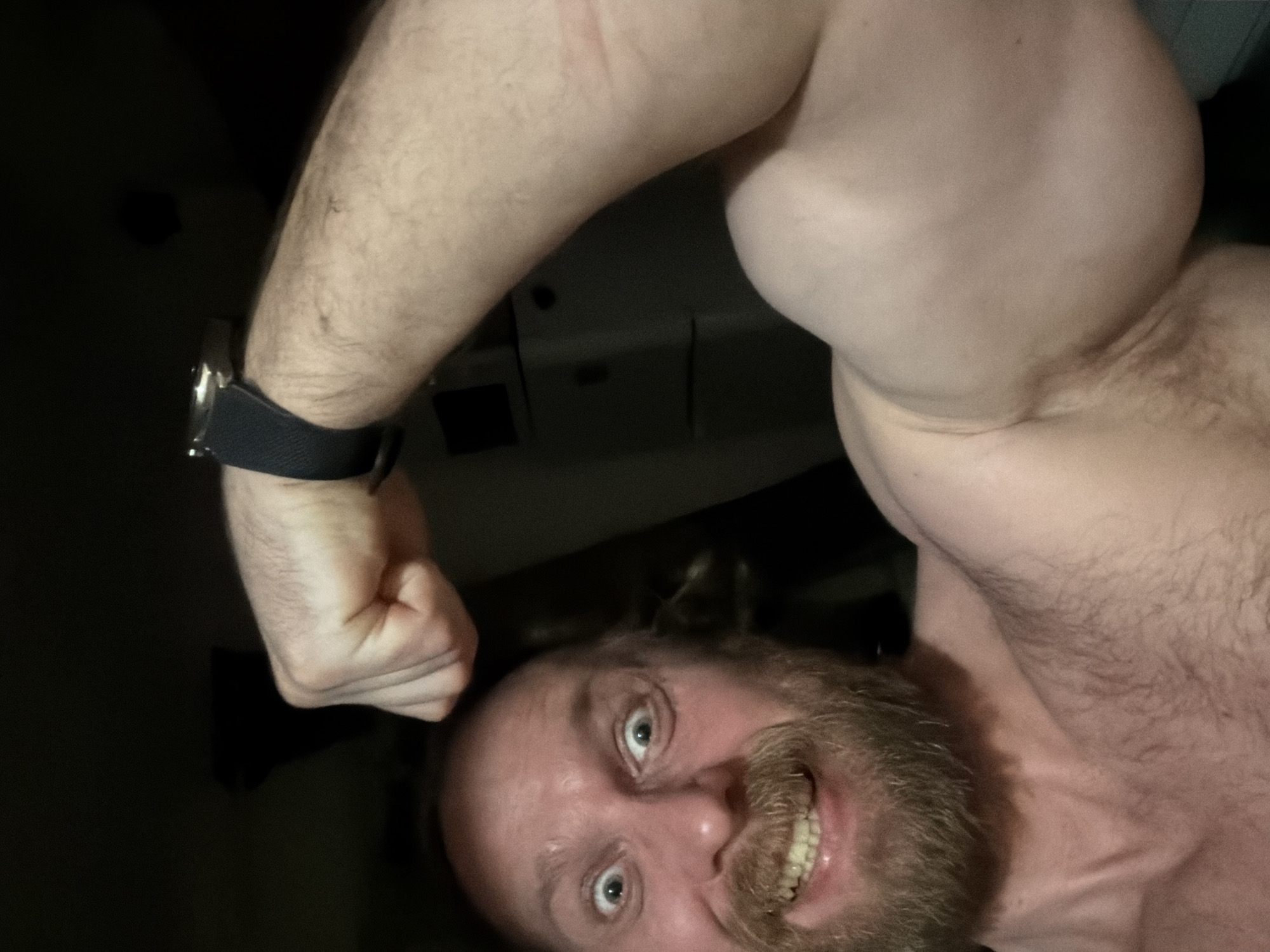 An adult man is depicted in a close-up, low-angle selfie, appearing inverted due to the camera's orientation. He has light skin, a full reddish-brown beard and mustache, and short or receding hair. His blue-green eyes are wide open, looking directly at the camera, and he displays a wide, toothy grin with his lower teeth visible, conveying an energetic or playful emotion.
The man is shirtless, showcasing his upper body. His right arm is prominently featured, flexed to highlight a muscular bicep. A watch with a dark strap and metallic casing is visible on his left wrist. The background is extremely dark and indistinct, suggesting an indoor setting with minimal lighting. No specific objects or features are discernible in the background. No visible text is present in the image. The provided location context is Saint Petersburg, Russia, though the image itself offers no visual cues to confirm this.