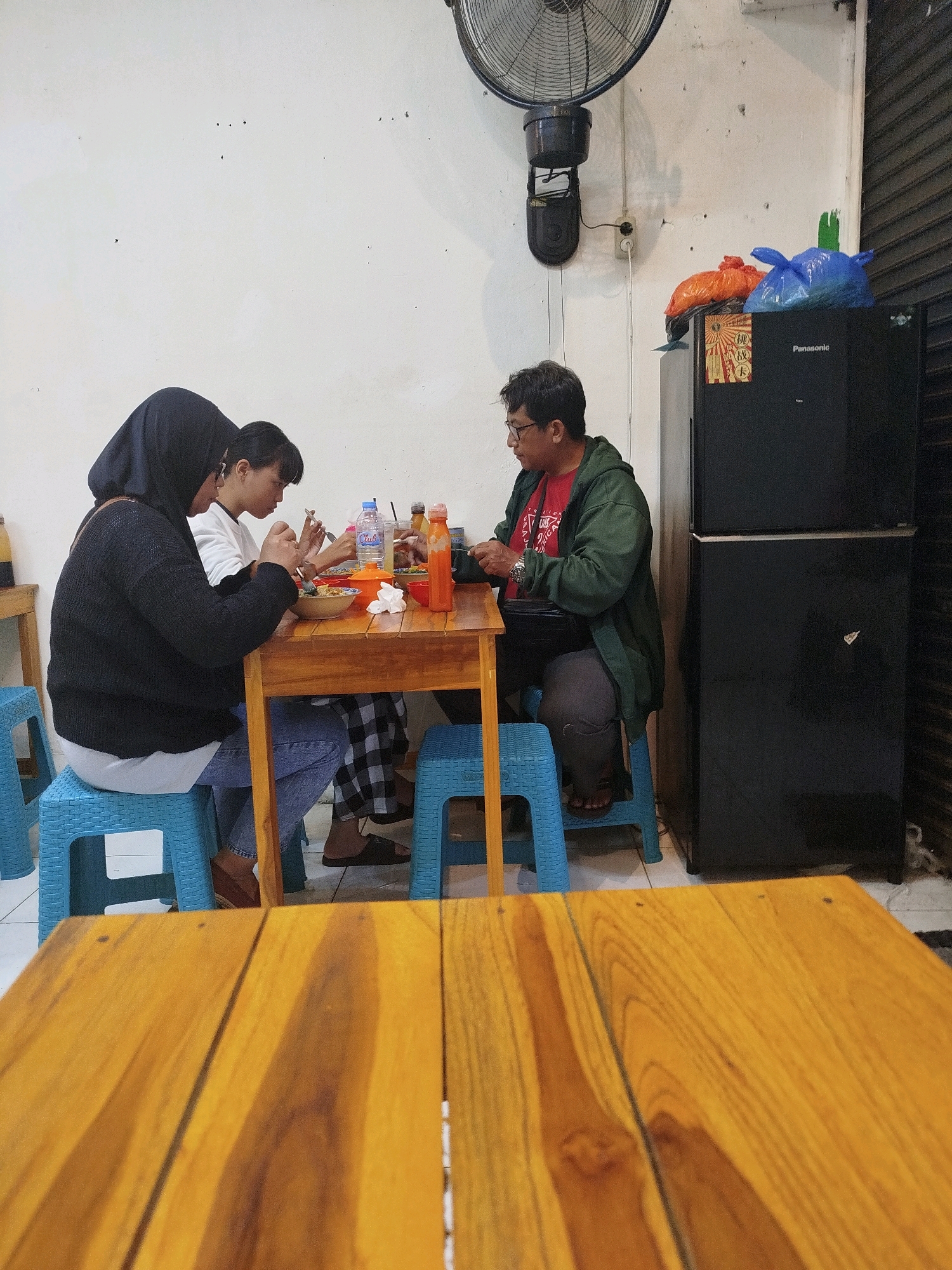 In a casual eatery in Surabaya, Indonesia, three individuals are seated around a wooden table, engaged in eating. The scene takes place indoors with white walls and a large fan mounted on the ceiling. A black Panasonic refrigerator stands against the wall on the right, with two plastic bags, one orange and one blue, resting on top.
The person on the left is a woman wearing a black hijab and a dark sweater over a lighter garment. She is leaning forward, eating from a bowl with a spoon. To her right sits a younger woman in a white top and checkered pants, also eating from a bowl with cutlery. Across from them, a man with glasses, wearing a red t-shirt under a green jacket, is also eating, holding a spoon. On the table are bowls of food, bottles of drinks, and condiments. The people are seated on blue plastic chairs. In the foreground, a wooden table surface is visible, slightly out of focus.