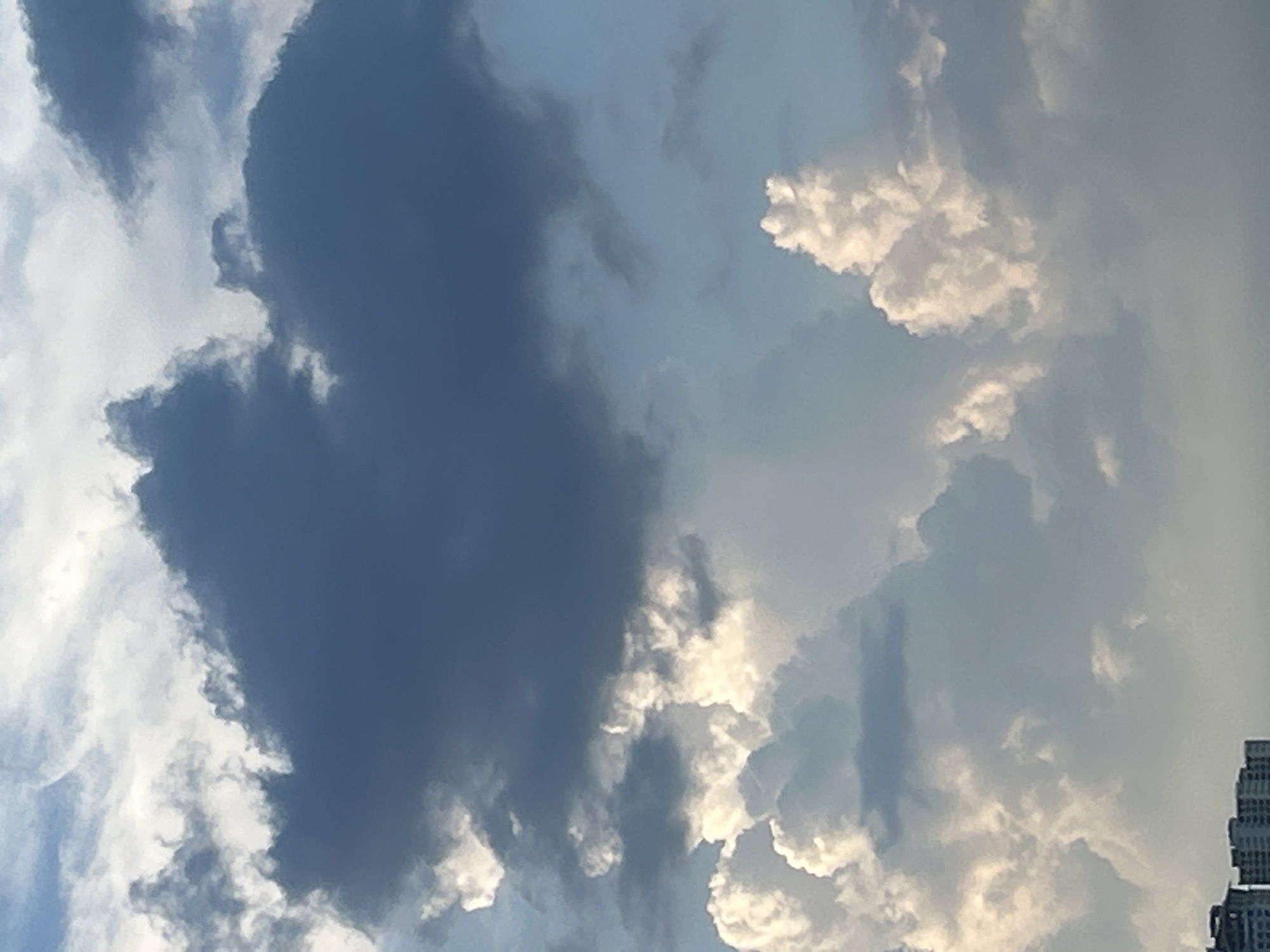 The image depicts a cloudy sky with hints of buildings at the bottom. The clouds are varied in texture and color, ranging from dark gray to bright white. Some clouds appear heavy and low, while others are wispy and high. The sky is a pale blue, with light filtering through the clouds. The buildings in the foreground are partially obscured by the clouds and appear to be apartment or office buildings, typical of an urban environment. The overall impression is one of a dynamic sky over a city.