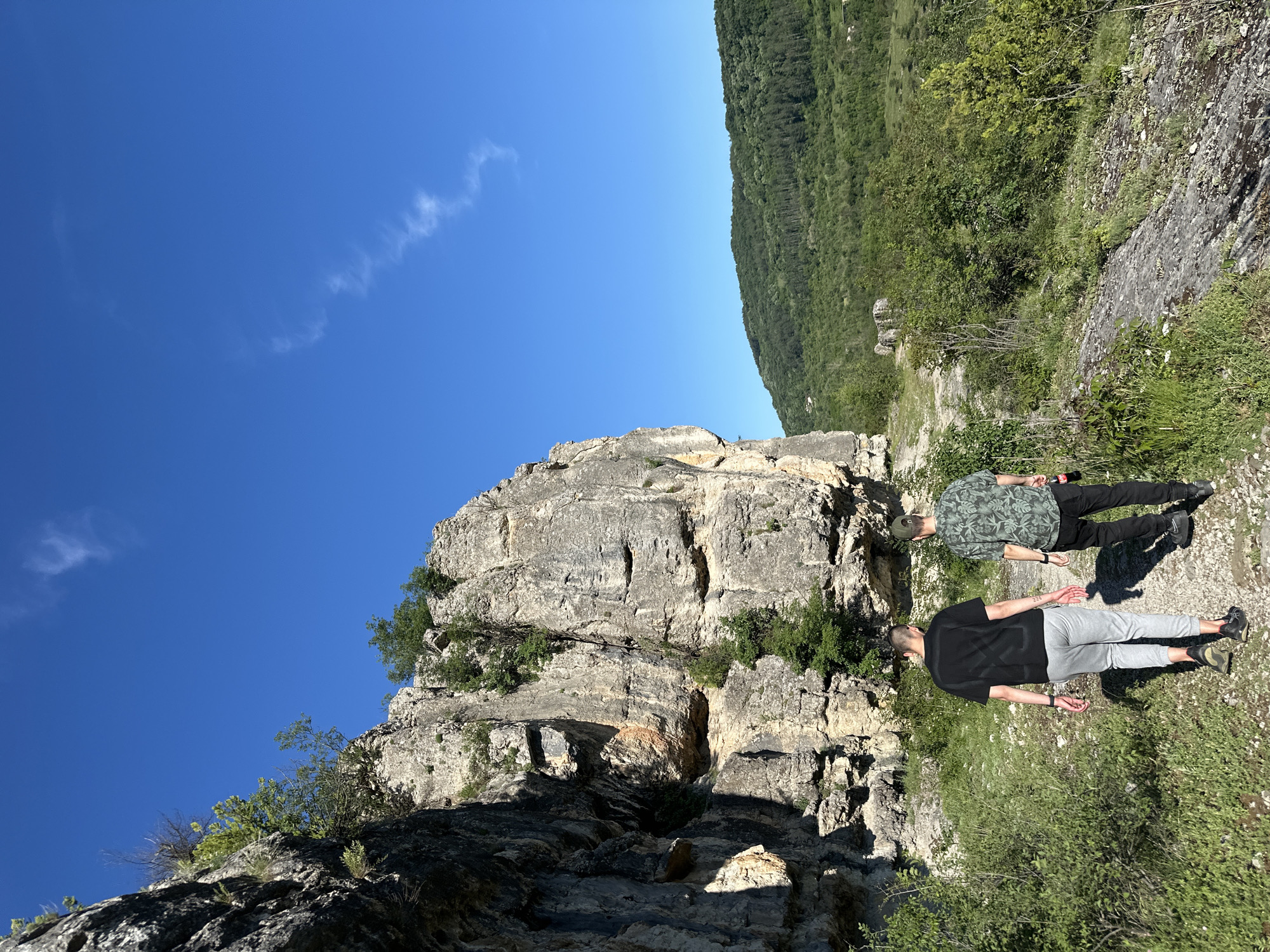 The image depicts a scene near a rocky cliff in Lovech, Bulgaria. Two men are lying on the ground. One man wears a green patterned shirt and black pants; the other wears a black t-shirt and grey pants. The cliff face is large and grey with some greenery growing on it. A forest is visible in the background, along with a clear blue sky with a wisp of cloud. The ground near the men is covered with green vegetation.