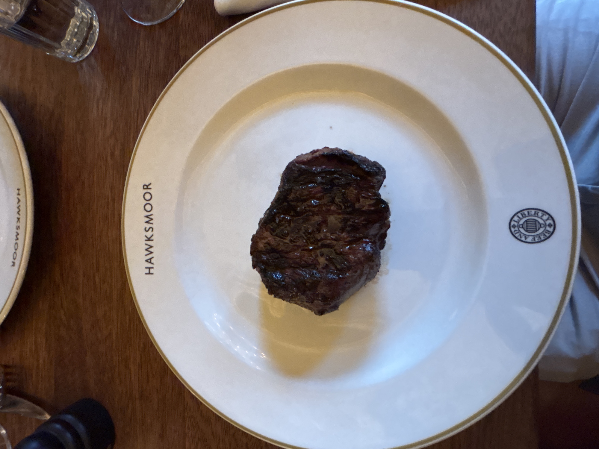 The image captures a close-up shot of a steak served on a white plate with a gold rim. The plate features the branding of Hawksmoor, a steakhouse chain. The writing "HAWKSMOOR" is on the left of the plate, and the logo of "LIBERTY AND BEEF" is on the right side. The steak appears to be well-cooked, with visible grill marks and a dark brown color. The plate is placed on a dark wooden table. Parts of a water glass and a second plate are also visible in the frame. The edge of a person's clothing is present on the right-hand side of the plate. Since the picture features a Hawksmoor branded plate, it can be inferred that the photo was taken inside a Hawksmoor restaurant, though a specific location in London cannot be determined with the information available.