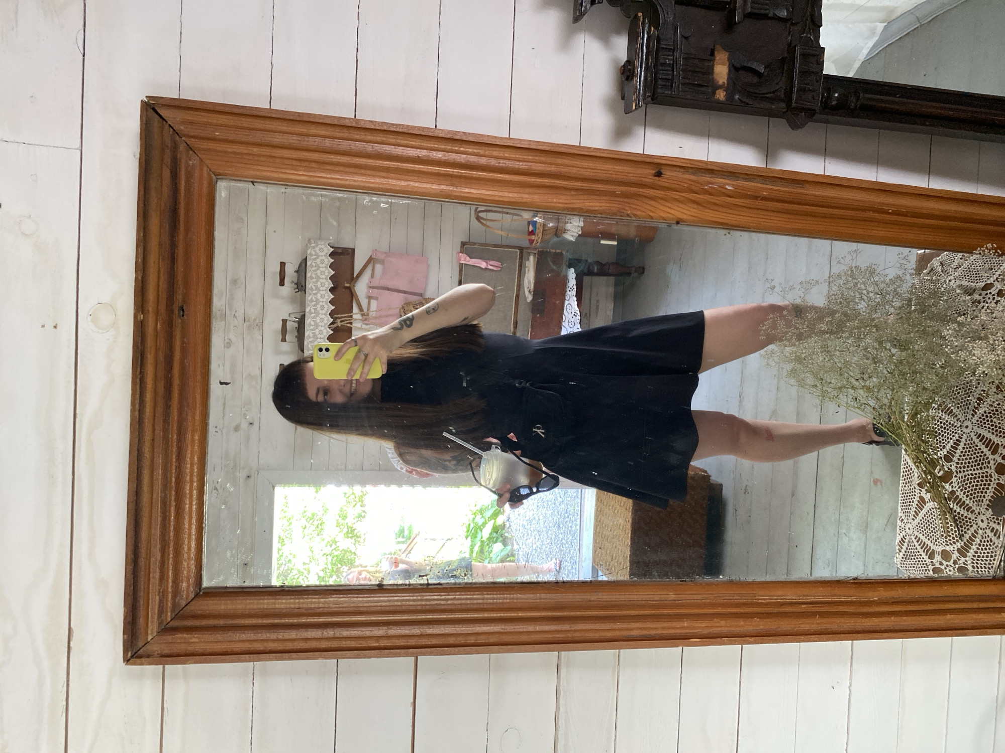 A young woman is taking a selfie in a large, wooden-framed mirror. She is wearing a black dress and holding a drink and sunglasses. The mirror reflects a room with white wooden walls, a doorway to the outside, and various antique items. It is difficult to determine the exact location in Votkinsk, Russia, but the rustic setting suggests a historical or vintage-themed establishment.
