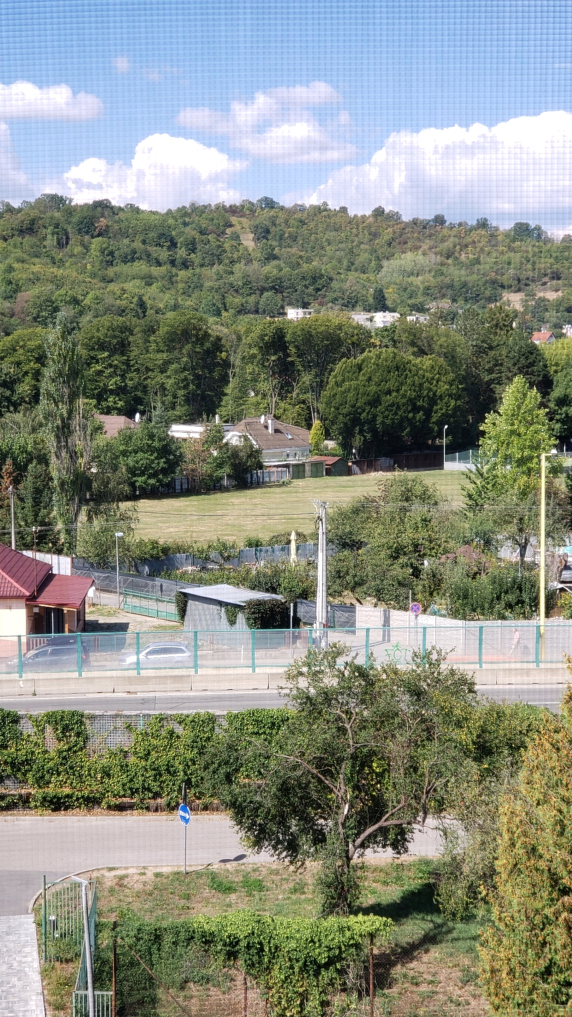 This is an exterior, daytime, high-angle shot of a suburban landscape. In the foreground, a street runs horizontally across the bottom of the image, bordered by a paved walkway and dense green hedges. A tall, slender tree with a sprawling canopy stands in the middle ground, casting a long shadow. Beyond the street, a fence separates it from a property with more hedges and a paved area with a blue directional sign. Further back, a metal fence encloses a property with a corrugated metal shed and a building with a red-tiled roof. Several vehicles are visible parked behind the fence. The middle ground also features a large, open, grassy field, bordered by trees and several residential houses with peaked roofs. In the background, a densely wooded hill rises, with some buildings visible through the trees. The sky above is blue with scattered white clouds. The scene appears to be located in Prešov, Slovakia, based on the provided context.