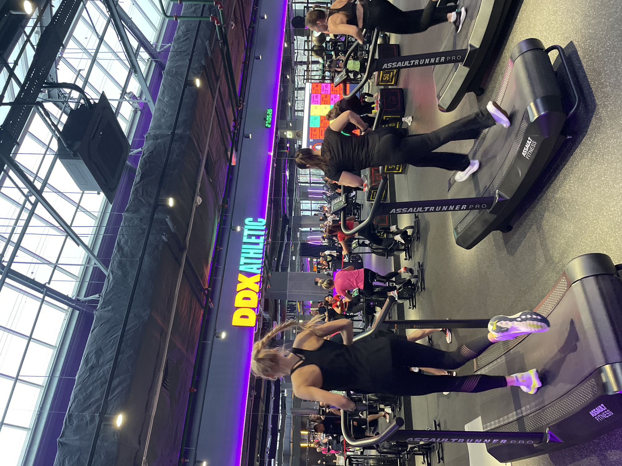 The image shows the interior of a gym, likely in Kazan, Russia, due to the Cyrillic lettering in the timestamp "27.05.26" (May 27, 2026).
The foreground features several curved, manually-powered treadmills labeled "ASSAULT RUNNER PRO" and "ASSAULT FITNESS." Individuals are actively using these treadmills, engaged in running or jogging.
In the middle ground, there are more treadmills and stationary bikes, with people using them. Further back, more gym-goers are visible.
The gym has a high ceiling with visible support beams and a translucent roof, allowing natural light to enter. On the left, a wall is adorned with the brand name "DDX ATHLETIC" in yellow and blue lettering, illuminated by a purple light strip running vertically. There are air conditioning units and wrapped ductwork visible near the ceiling.