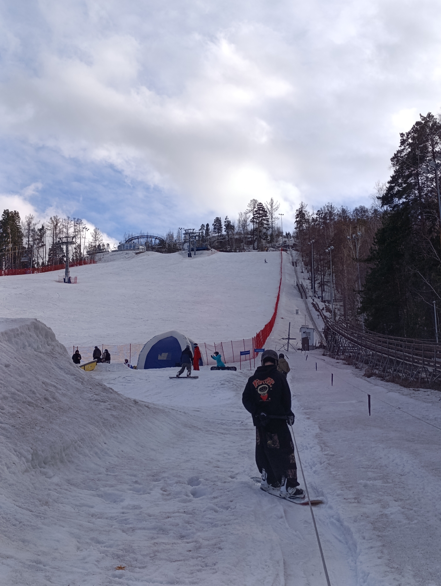 The image depicts a winter scene at a ski resort in Krasnoyarsk, Russia. In the foreground, a person on a snowboard is being pulled uphill by a rope tow. They are wearing a black hoodie with a graphic on the back, camouflage pants, and white boots. The person is also wearing gloves. A wooden walkway runs along the right side of the frame, bordering the rope tow.
In the midground, a ski slope is visible with several people scattered across it. There is a blue and white tent-like structure near the bottom of the slope. Further up the slope, there are red safety fences and several ski lifts.
The background features a snow-covered hill with trees and a cloudy sky. The buildings and infrastructure of the ski resort are visible at the top of the hill.