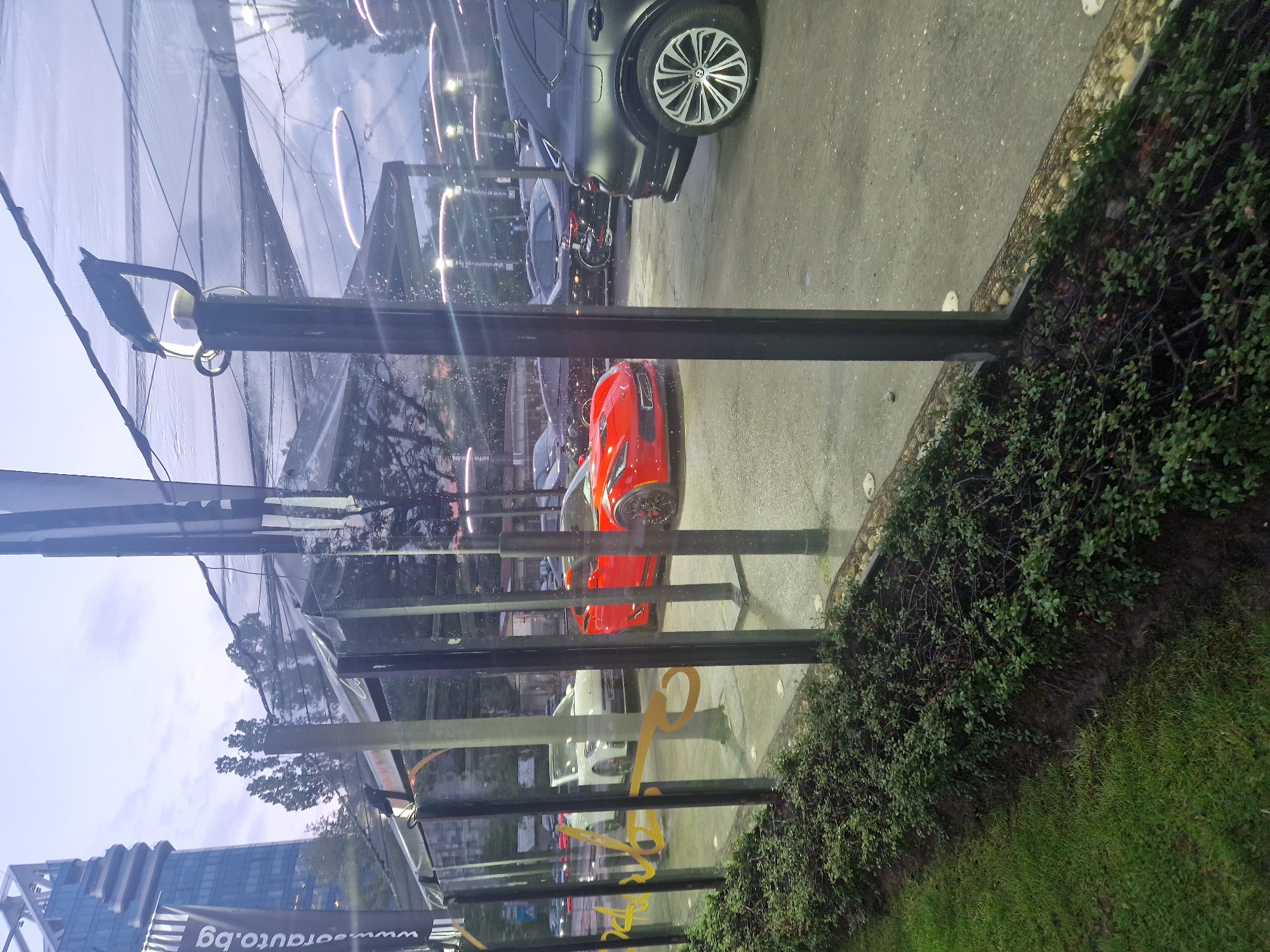 The image shows a car dealership or showroom viewed through a glass facade in an urban environment. A bright red sports car is parked prominently, followed by other vehicles including a black SUV with distinctive wheels. The setting includes greenery like a hedge or bushes near the glass exterior. Reflections of lights are visible on the glass. A banner with the URL "www.arauto.bg" is partially seen. The location is in the city of Unknown, Unknown.