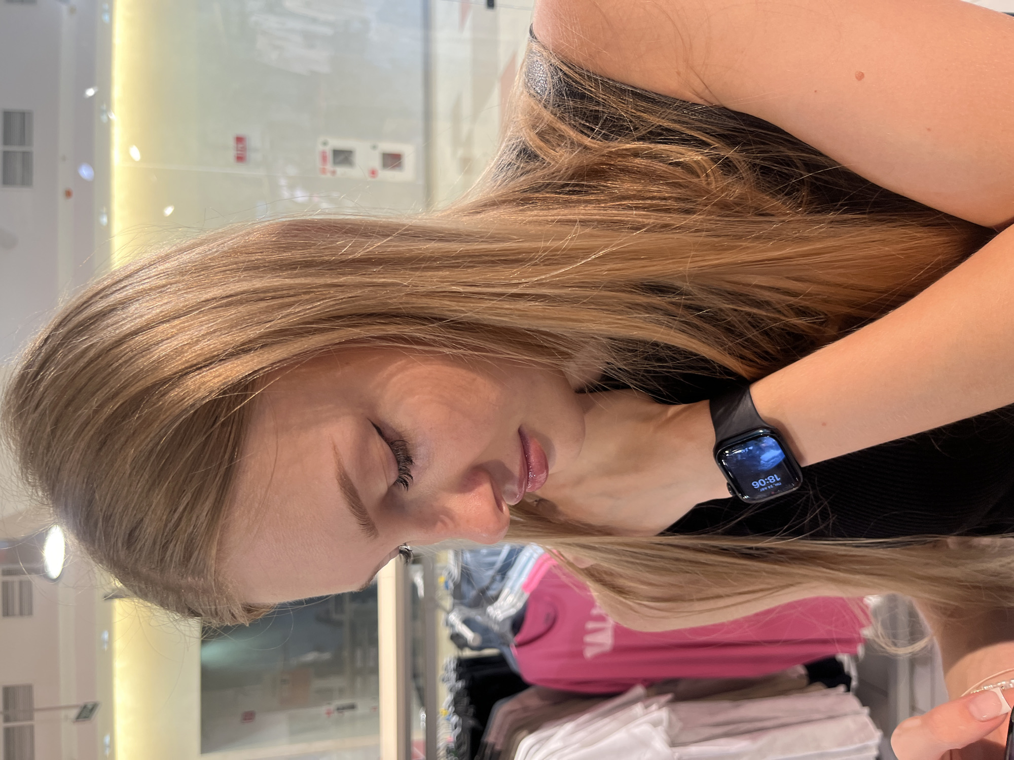 A close-up shot shows a young woman looking down at her wrist where she is wearing a smartwatch. The woman has long, blonde hair and is wearing a black top. She is in a retail environment, with racks of clothing visible in the background. The scene appears to be well-lit with artificial lighting. The image does not provide any specific information or visual cues to determine the location within the city of Perm, Russia.