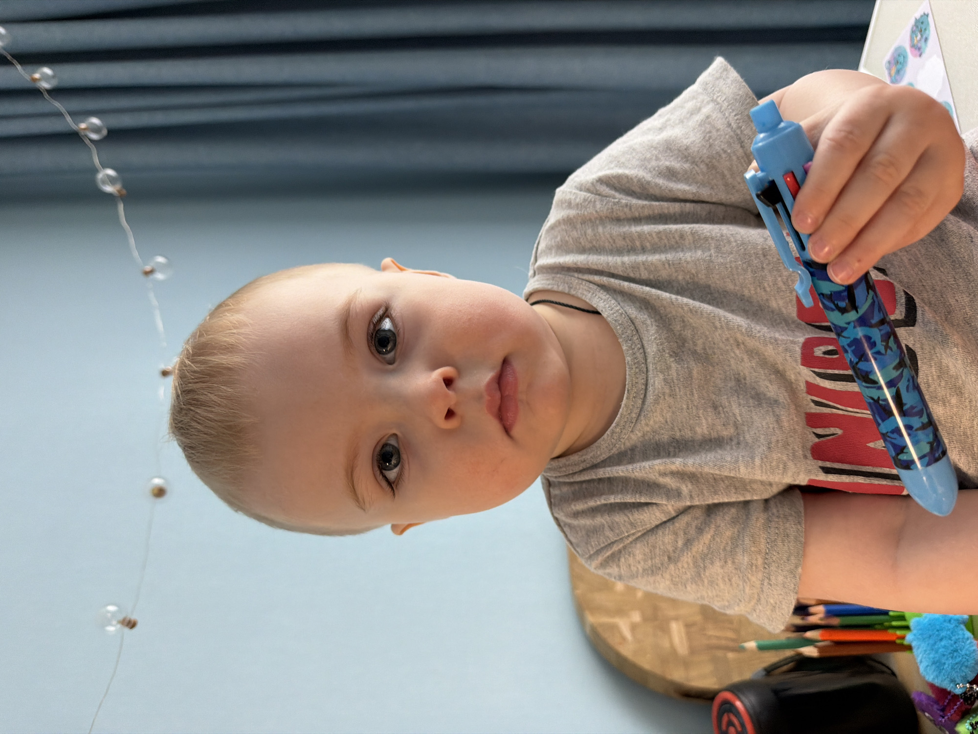 A young, light-skinned child with blonde hair holds a blue multi-color pen in their right hand. The child is wearing a gray t-shirt with red and black details. The background includes a light blue wall, decorative string lights, and a glimpse of what appears to be a table with colored pencils, a black case, and a blue pompom. The image is rotated counter-clockwise. The location is unknown, however the user has requested that it be located in the city of Votkinsk, Russia.