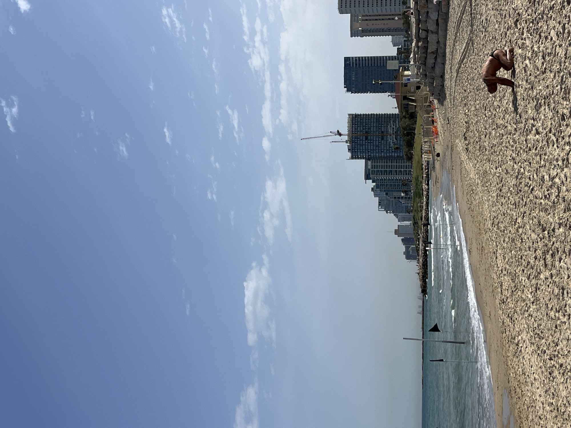 A beach scene in Tel Aviv, Israel is presented from a slightly elevated perspective. Along the shoreline, a man clad in dark swimwear is positioned on all fours, facing towards the sea. Parallel to the beach, tall buildings constitute the city skyline. Between the sand and the buildings lies a row of vegetation. A crane is visible amidst the buildings, suggesting construction activities. Flags stand in the water. The sky is clear with scattered clouds.