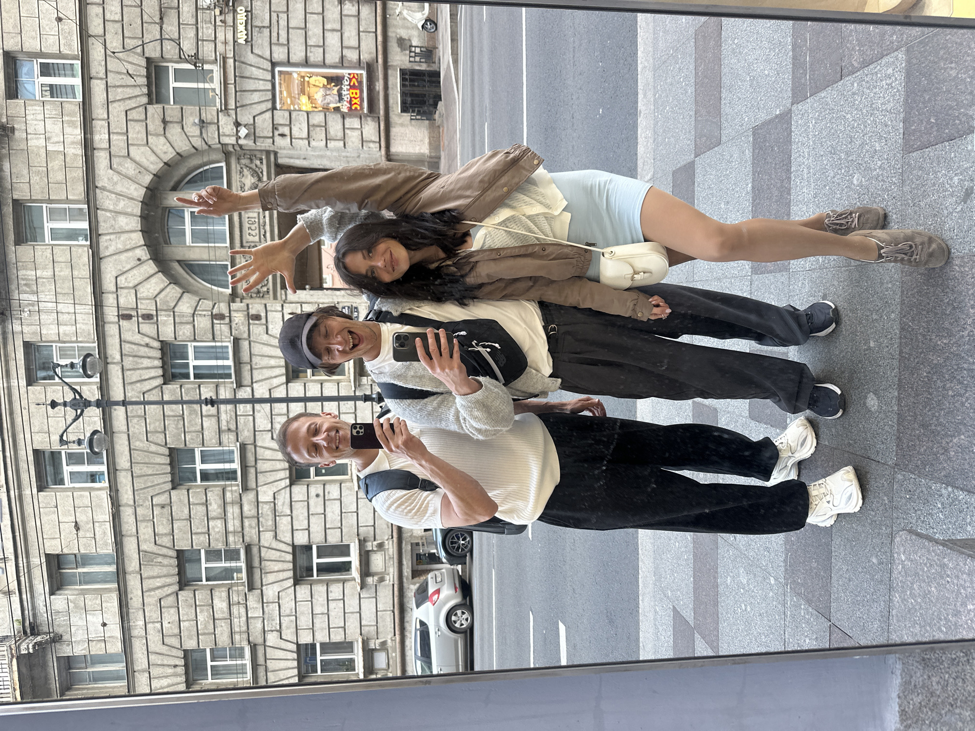 Three individuals, two men and one woman, are captured in a mirror selfie on a sidewalk in Saint Petersburg, Russia. The background features a classic St. Petersburg building with ornate stone facade and arched windows. A street lamp with multiple lights stands to the right of the individuals.
The man on the left wears a white ribbed polo shirt, black pants, and white sneakers, with a black backpack. He is holding a smartphone and smiling. The man in the middle wears a light grey cardigan over a white t-shirt, dark grey pants, and black sneakers, also holding a smartphone. He is laughing with his mouth open. The woman on the right is wearing a light blue dress, a cream-colored cardigan, and a brown jacket. She has a small white crossbody bag and is wearing brown sneakers. Her left arm is raised in the air with her index finger pointed upwards, and her right arm is raised with her hand open.
A silver car is parked on the street behind the individuals. The sidewalk is paved with granite tiles of varying grey tones. The overall lighting suggests it is daytime, likely overcast.