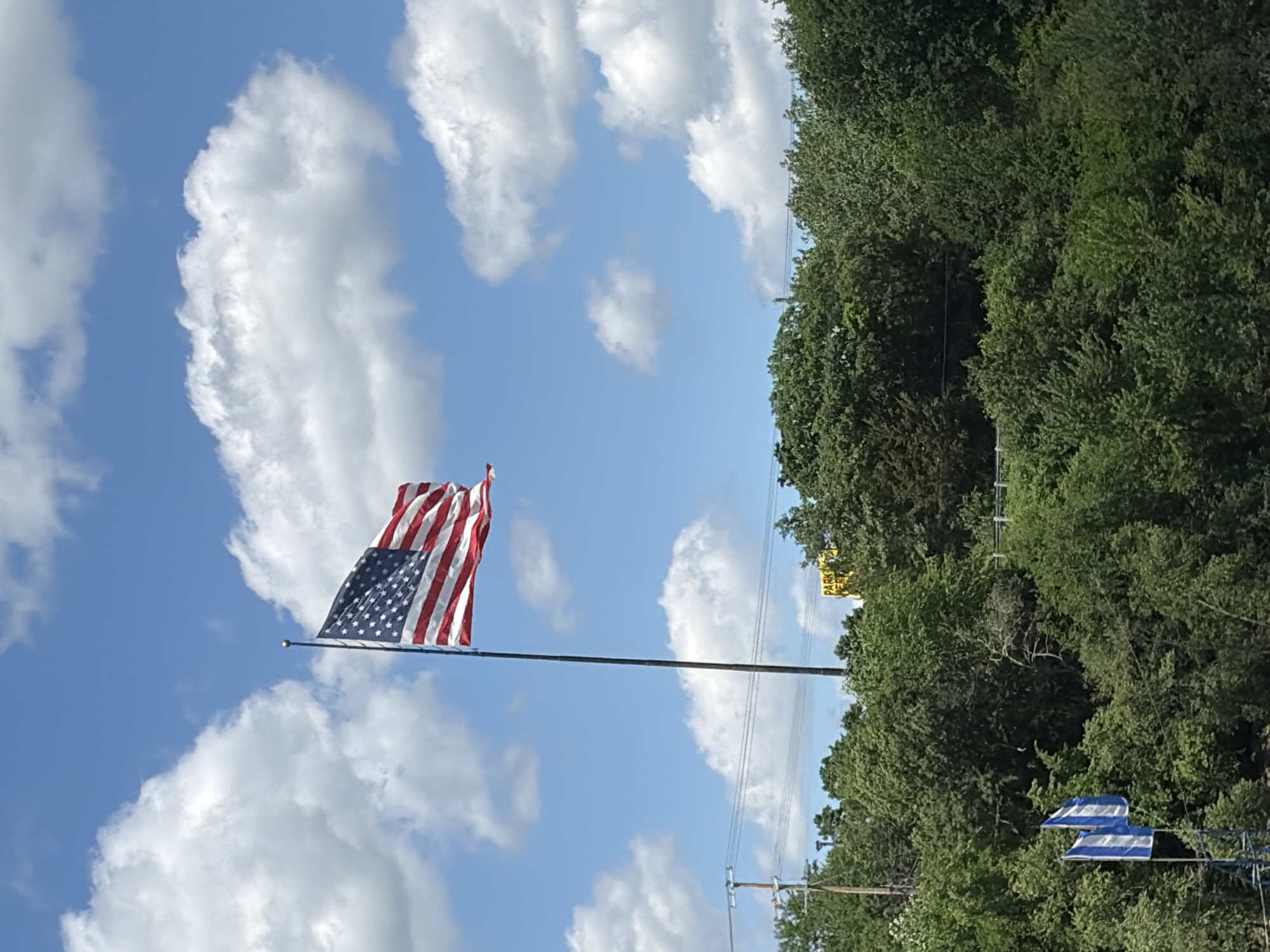 An American flag flies at half-mast from a tall flagpole against a bright blue sky dotted with fluffy white clouds. The flag is positioned in the upper left quadrant of the image. Below the flag, a dense canopy of lush green trees fills the lower portion of the frame, with some taller trees reaching towards the sky. In the distance, utility poles and power lines are visible, suggesting an urban or suburban setting. A yellow sign with the word "PARK" is partially obscured by trees on the right side of the image. Two smaller flags, one with blue and white stripes, are visible on the left, planted on a shorter pole. The overall scene captures a patriotic display set against a natural backdrop on a sunny day. The scene is likely located in Wisconsin Dells, United States of America.