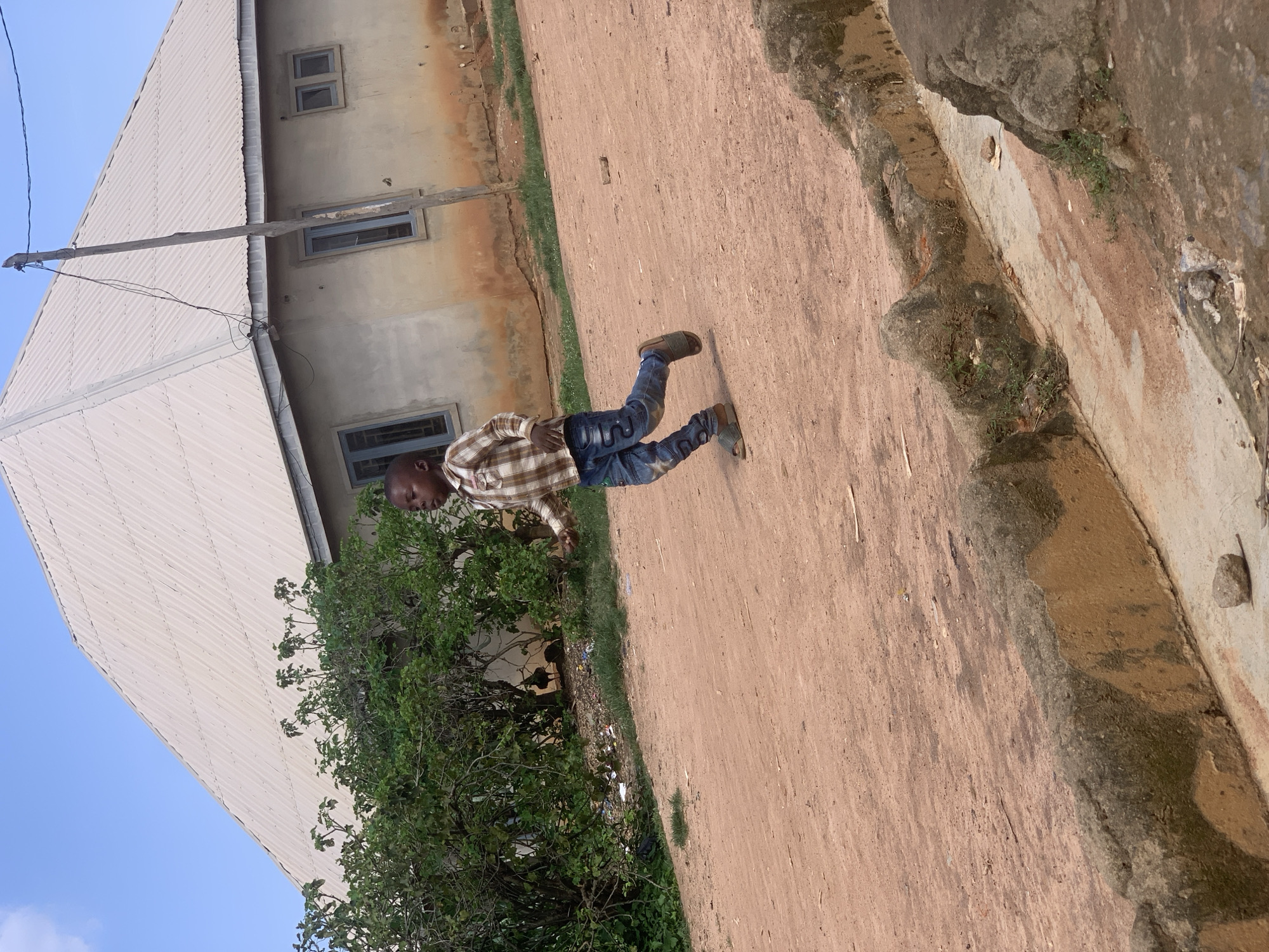 In Sabon Tasha, Nigeria, a young boy is captured mid-stride, running across a dirt path. He wears a checkered long-sleeved shirt and blue jeans with a white star pattern. His feet are clad in dark-colored slip-on sandals. The boy is positioned on the right side of the frame, facing towards the left. Behind him, a pale grey, corrugated metal roof slopes down from a white house with grey-trimmed windows. To the left of the house, a dense green tree provides a backdrop. The ground in the foreground is a dusty, reddish-brown earth, with a weathered concrete structure running diagonally across the bottom portion of the image, bordered by sparse green vegetation. The sky above is a clear, pale blue.