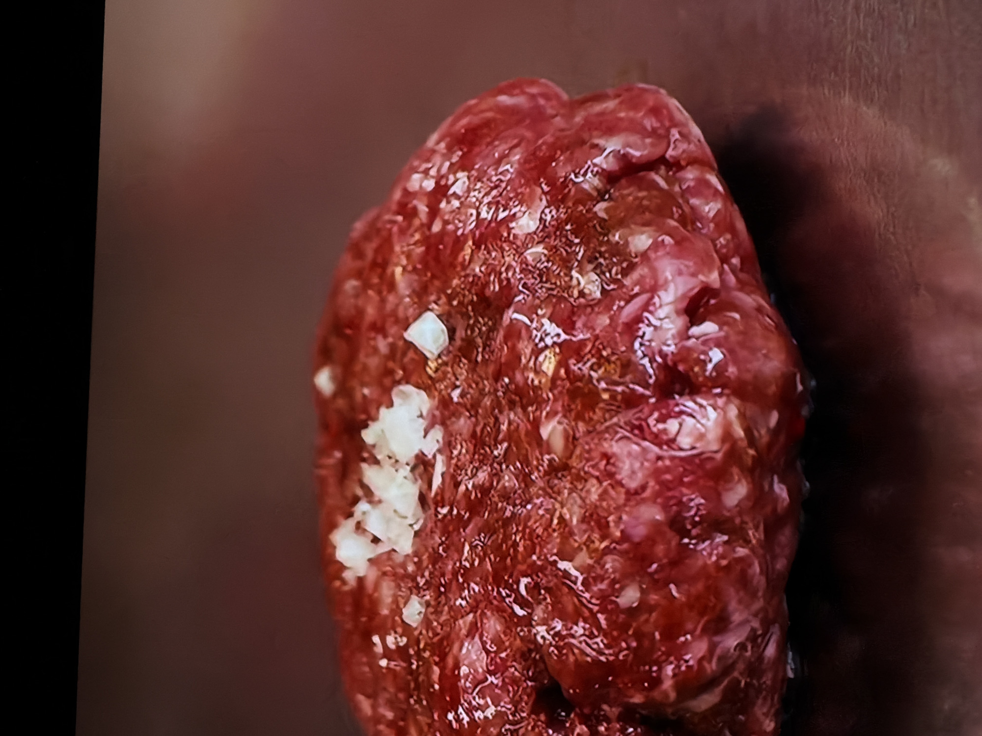 The image presents a close-up shot of a portion of raw, seasoned ground meat. The meat, likely beef or pork, displays a deep reddish-brown color and a coarse, somewhat lumpy texture. Scattered across its moist, glistening surface are small, irregular white pieces, which appear to be minced garlic, salt crystals, or finely chopped onions, indicating seasoning.
The meat is positioned on what appears to be a dark, possibly metallic or stone, surface. The background is out of focus, consisting of blurred shades of reddish-brown and dark gray, suggesting an indoor setting such as a kitchen countertop. A dark, undefined vertical band is visible along the far left edge of the frame. The lighting emphasizes the textured surface of the meat and the glistening moisture. There are no human subjects, discernible activities in progress, or visible text within the frame. The image focuses exclusively on the raw ingredient, highlighting its preparation.