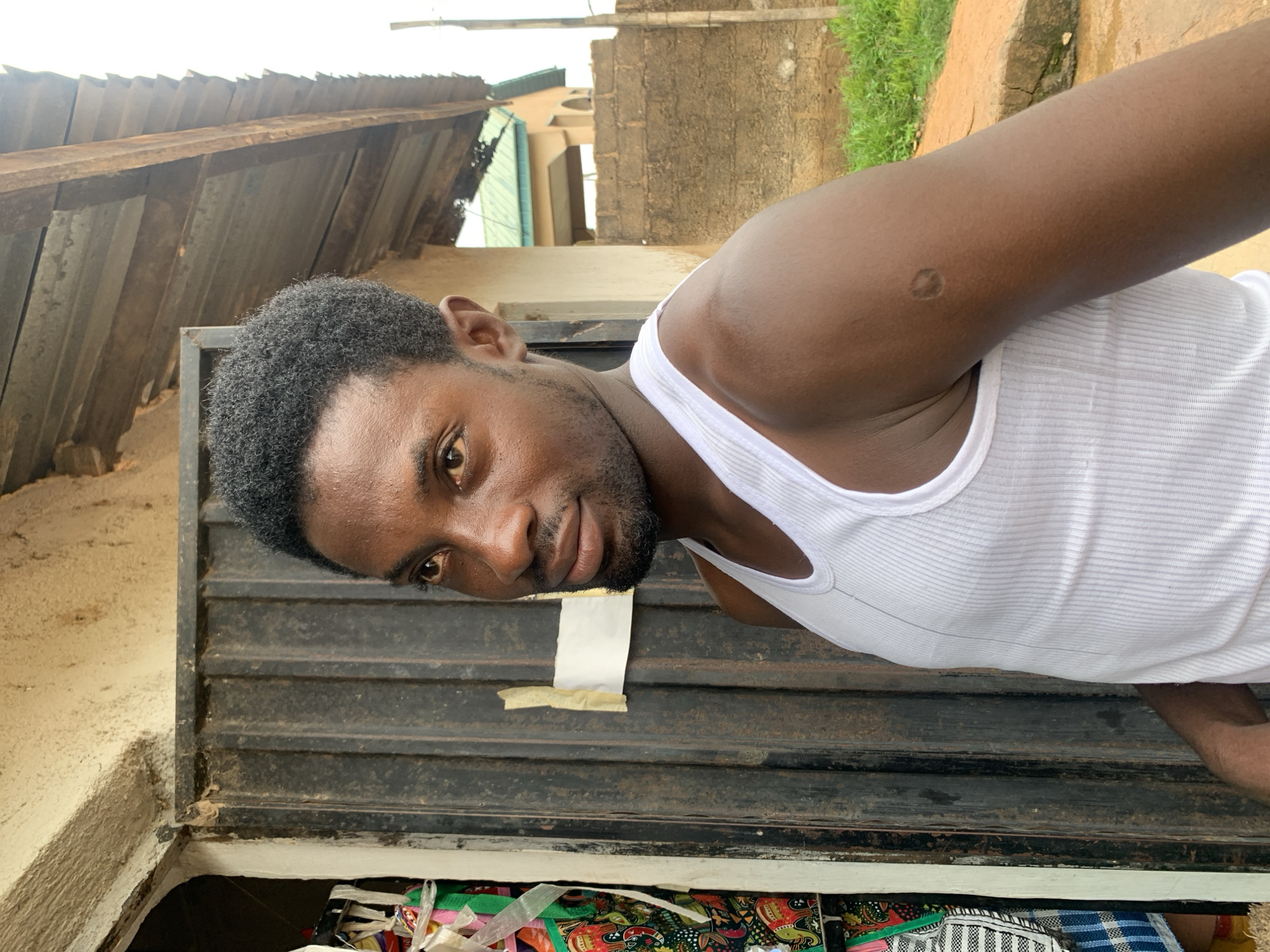 The media file shows a close-up, portrait-style, outdoor shot of a young Black man. He is wearing a white ribbed tank top and looking towards the camera with a slight smile. His skin is dark brown, and he has short, dark, curly hair and a beard. His left arm is resting on a dark metal surface that appears to be a rolling shutter door. There is a small, circular, lighter-colored mark on his upper left arm. The background is slightly blurred, showing some architectural elements that are consistent with the provided location context of Jos, Nigeria, including a corrugated metal roof and concrete walls. Some green foliage is also visible. The lighting suggests it is daytime, possibly late afternoon. The man's expression is relaxed and friendly. There is a piece of white paper taped to the metal door in front of him, but no visible text on it. Below the shutter door, a glimpse of colorful fabrics is visible, suggesting the area might be a shop or market stall.