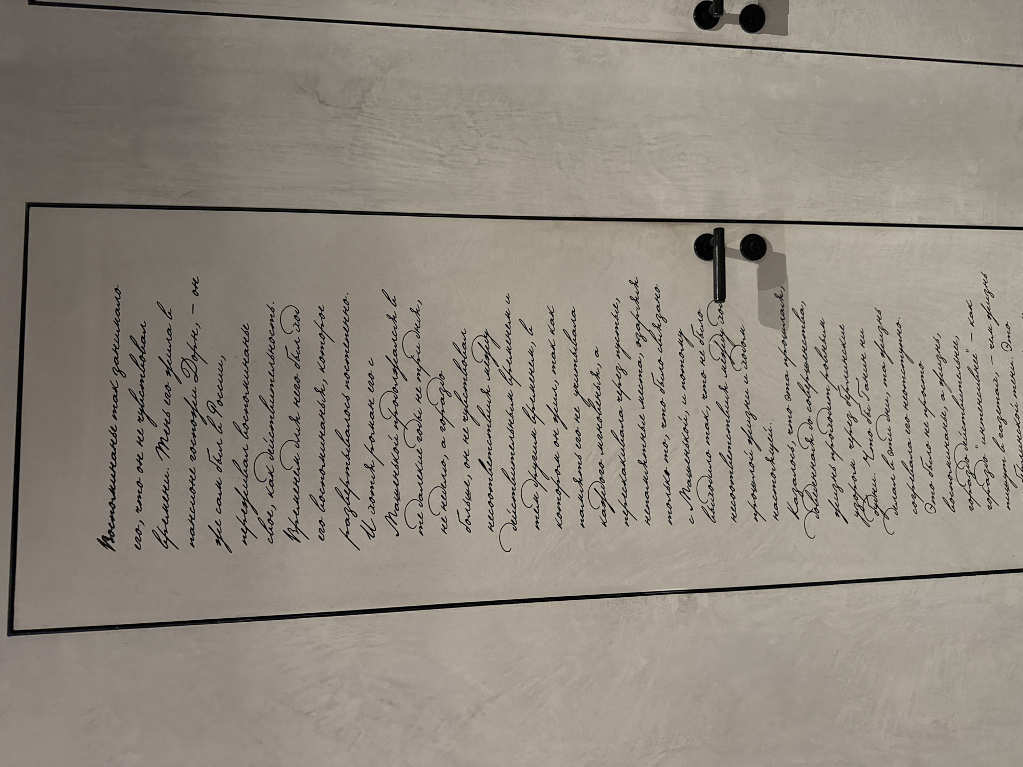 The image shows a section of a wall with handwritten text in Cyrillic script. The text is written in black ink on a light-colored, textured surface that appears to be part of an interior wall. The script is in a cursive style, making it somewhat difficult to read without close examination. There are two dark-colored door handles visible on the right side of the image, indicating this is likely a doorway or part of a room's interior. The handles are positioned at different heights on what appear to be two adjacent doors or panels. The text itself is densely written, covering a significant portion of the wall space. The overall scene suggests an artistic or decorative installation within a building. Without further context or visible landmarks, it is not possible to definitively determine the exact location within Moscow, Russia.
