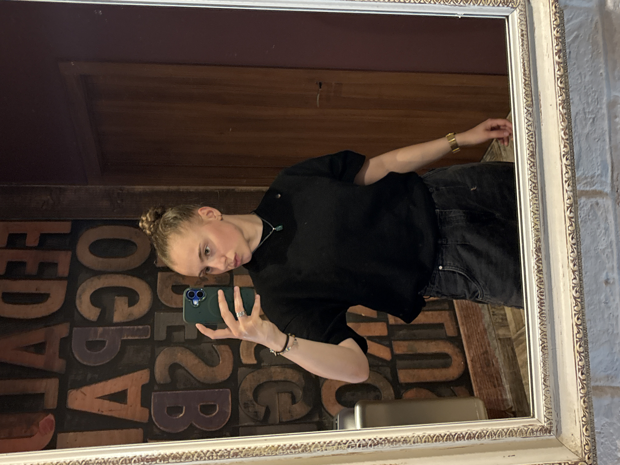 A young woman with her hair in a bun takes a mirror selfie in a room with a wooden door and a brick wall. She is wearing a black t-shirt and dark wash jeans. In her right hand, she holds a smartphone. She is wearing a gold watch on her left wrist and a necklace with a green pendant. The background of the room features a wall decorated with large, vintage letterpress type. The location is in Sofia, Bulgaria.